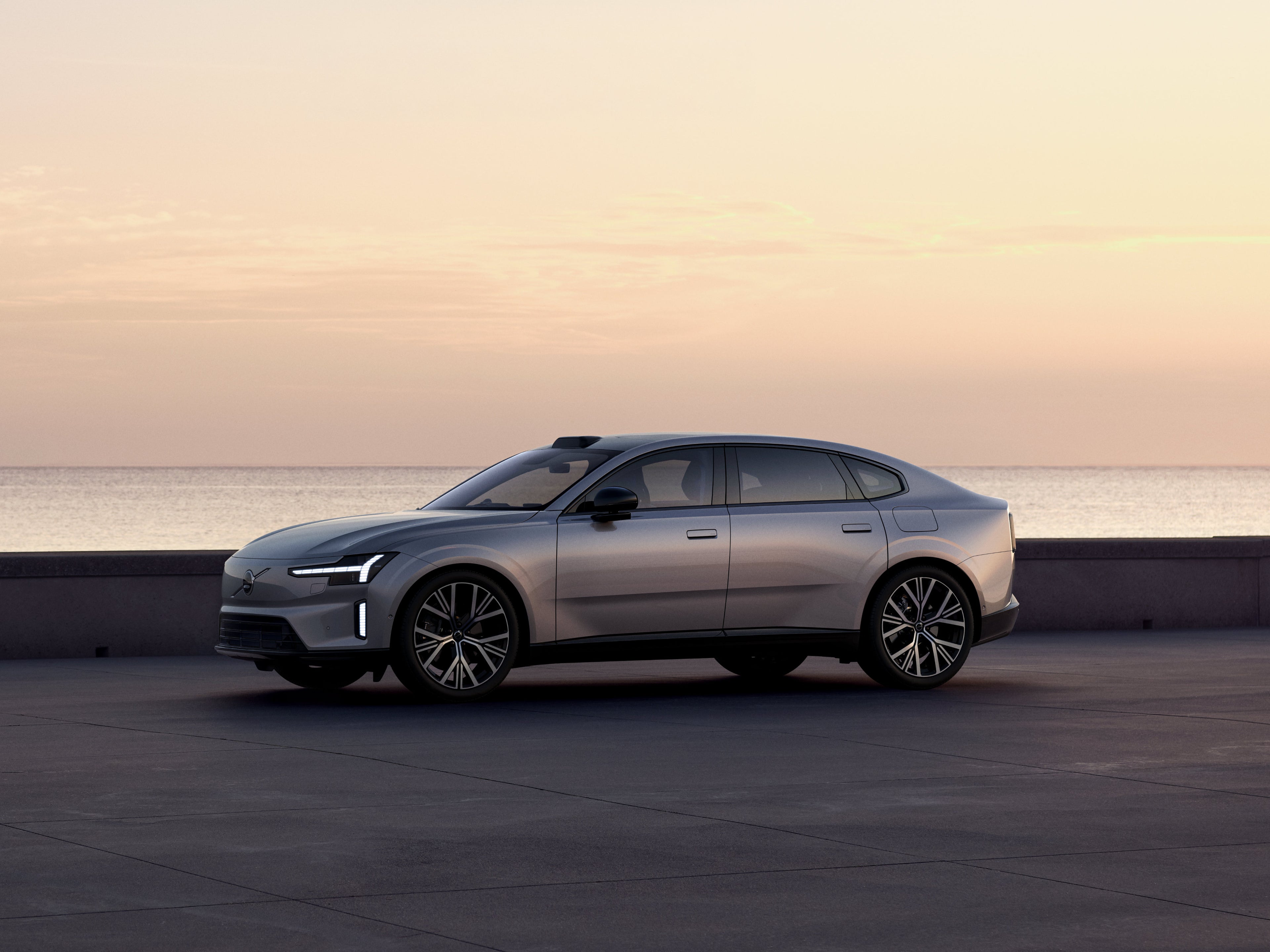 Ein silberner Volvo ES90 parkt bei Zwielicht an einem Küstenstreifen vor dem Meer.