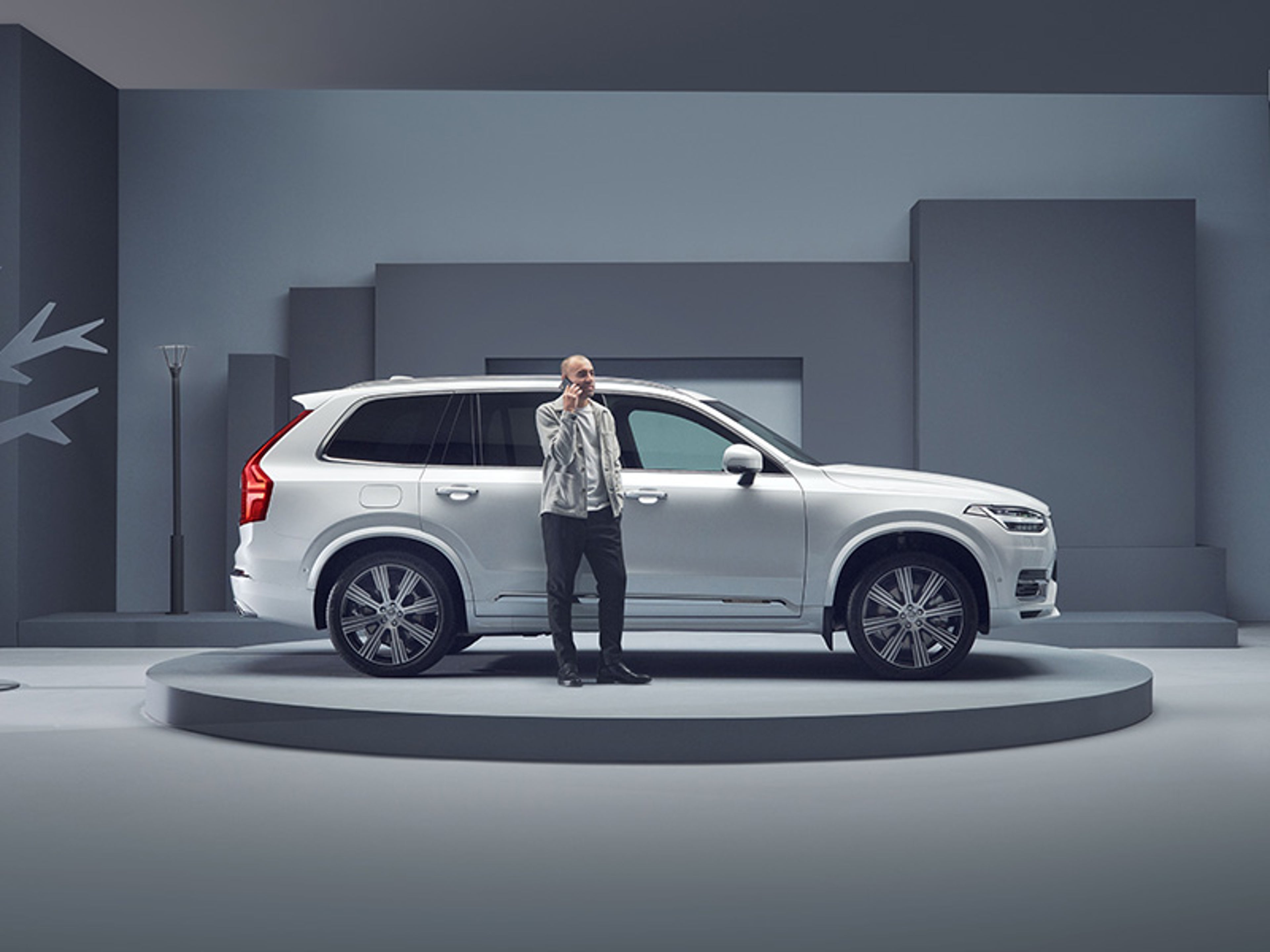 Un homme téléphone à côté d'une Volvo XC90 blanche sur un plateau tournant dans un studio gris.