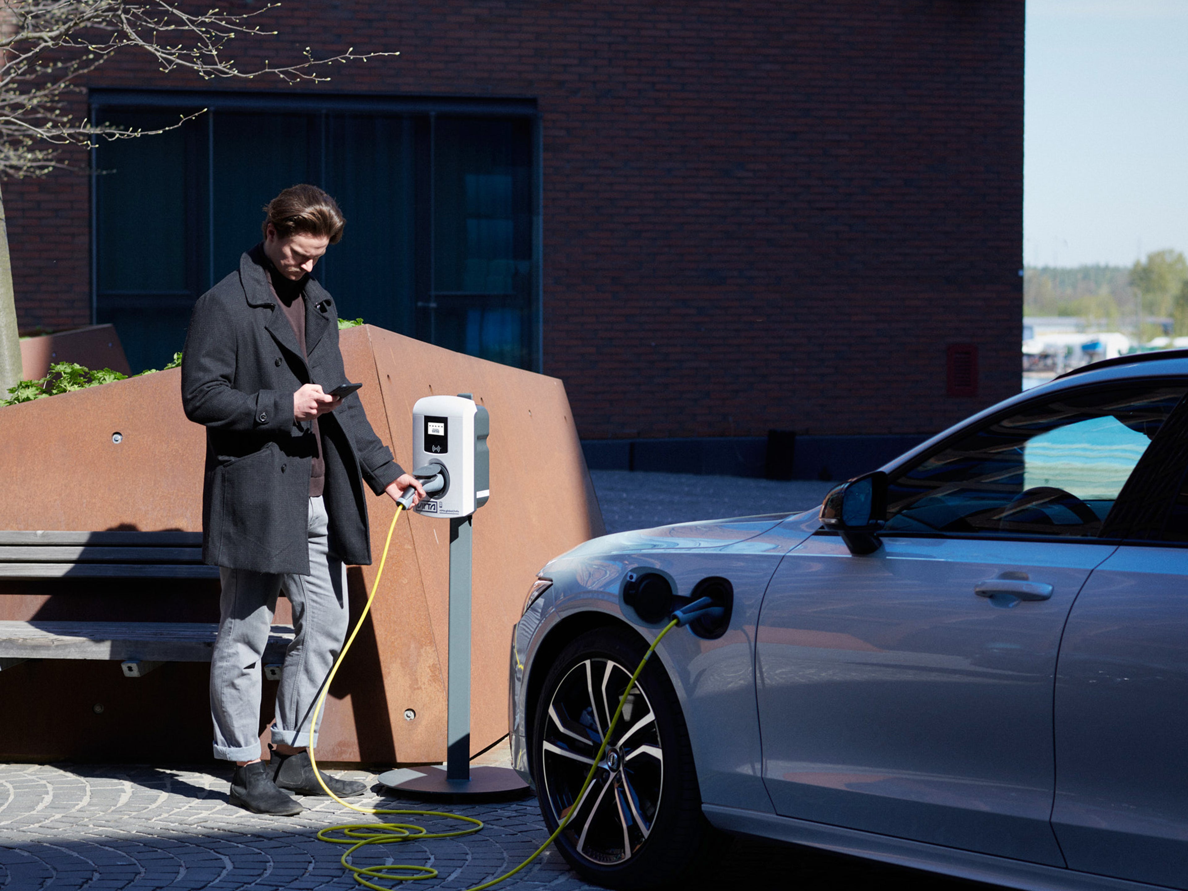 Mies seisoo Virta latausaseman ja lataukseen kytketyn Volvo auton vieressä ja seuraa latauksen edistymistä puhelimensa näytöltä.