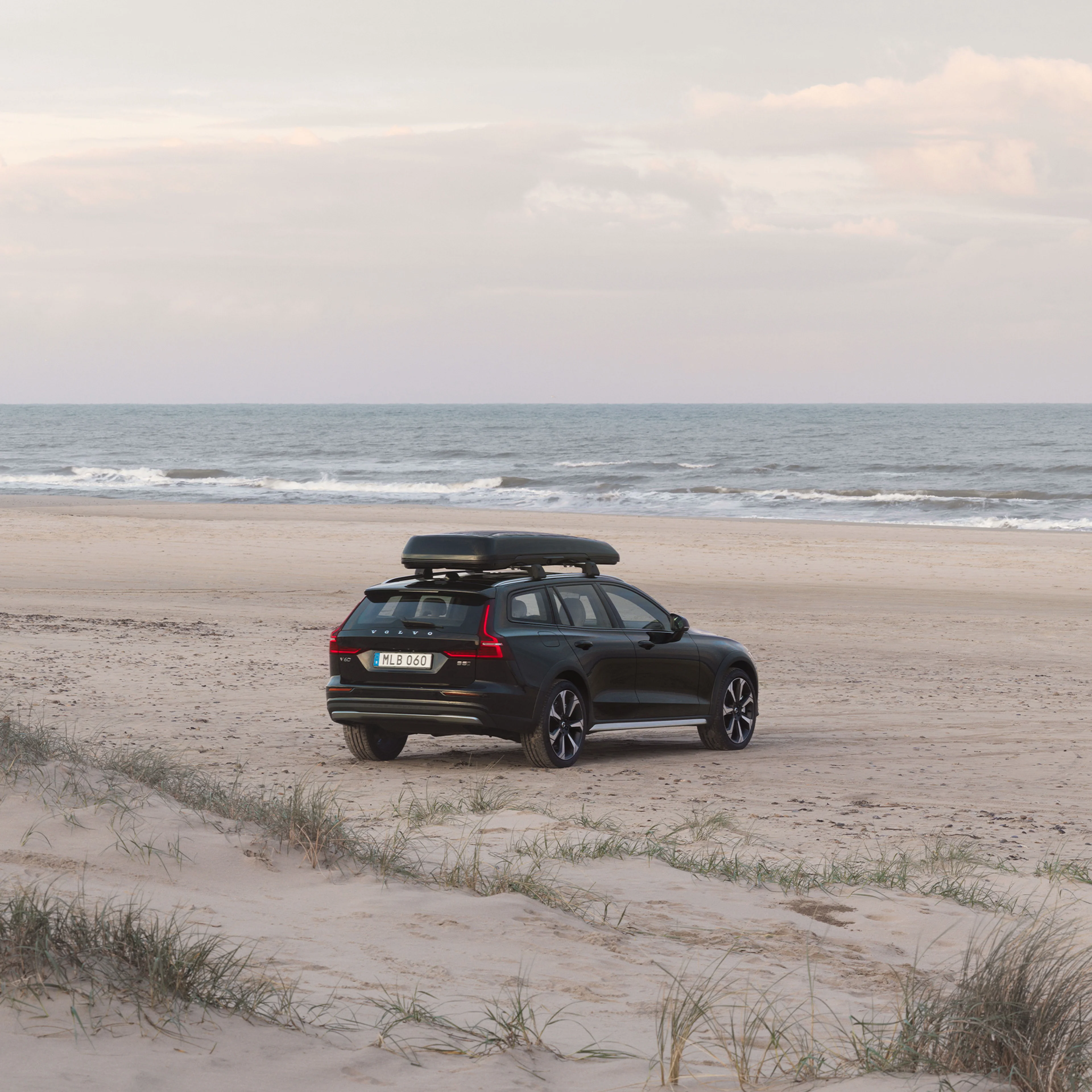 Ein schwarzer Volvo V60 Cross Country mit Dachbox steht auf einem Strand an der Küste mit der Fahrzeugfront zum Meer.