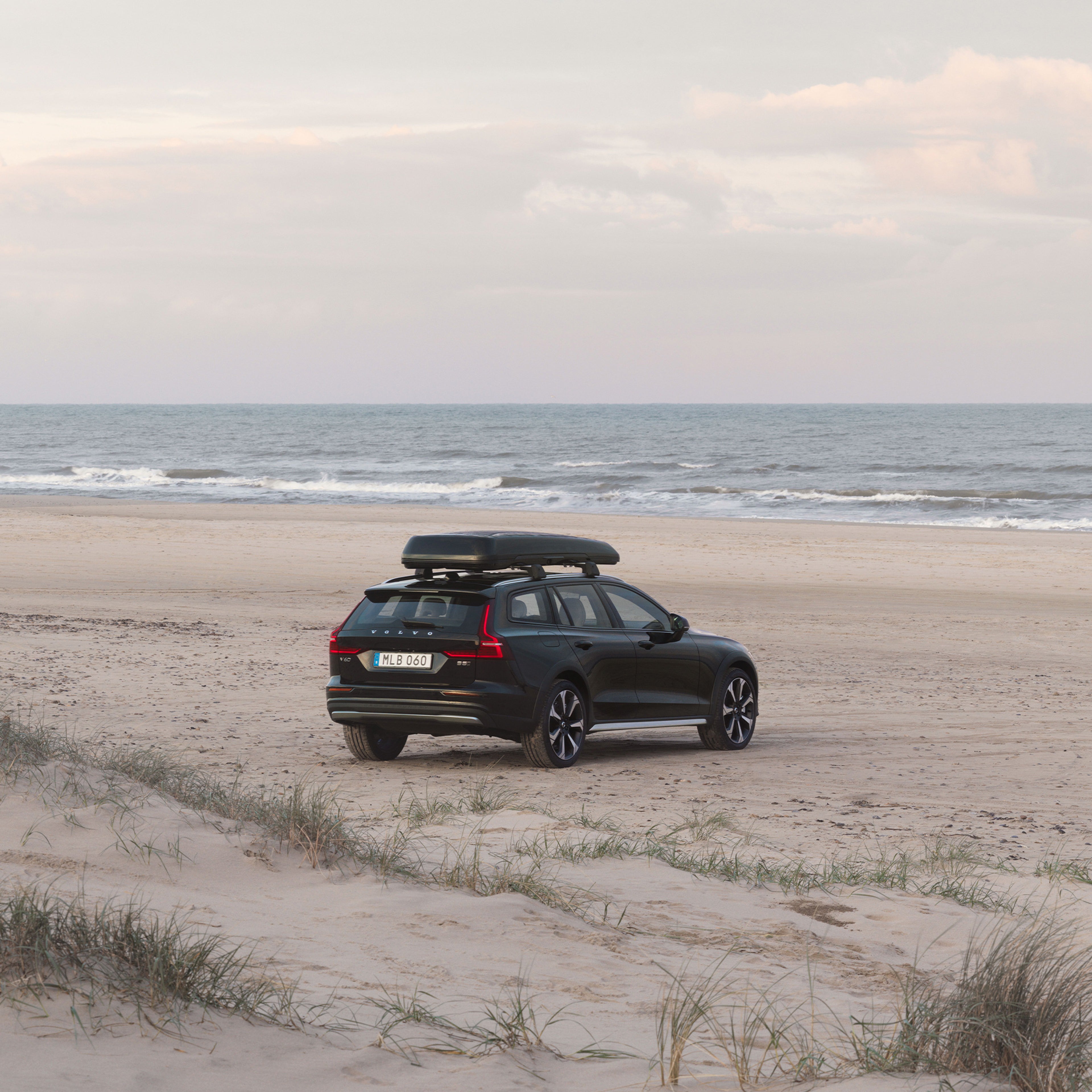 Ein schwarzer Volvo V60 Cross Country mit Dachbox steht auf einem Strand an der Küste mit der Fahrzeugfront zum Meer.