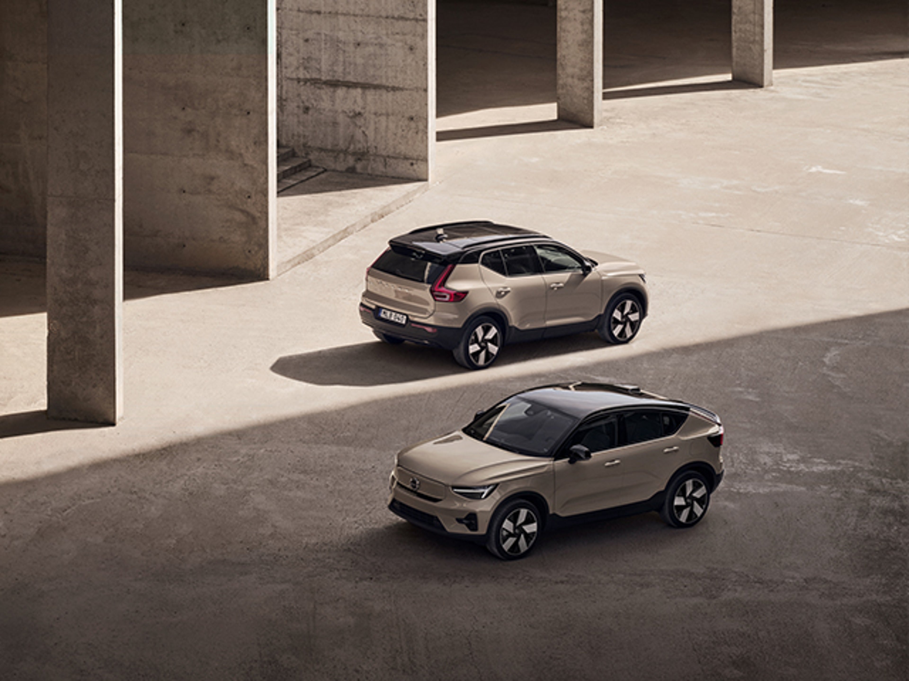 En sandfärgad Volvo EC40 och en sandfärgad Volvo EX40 står parallelt med varandra i betongmiljö