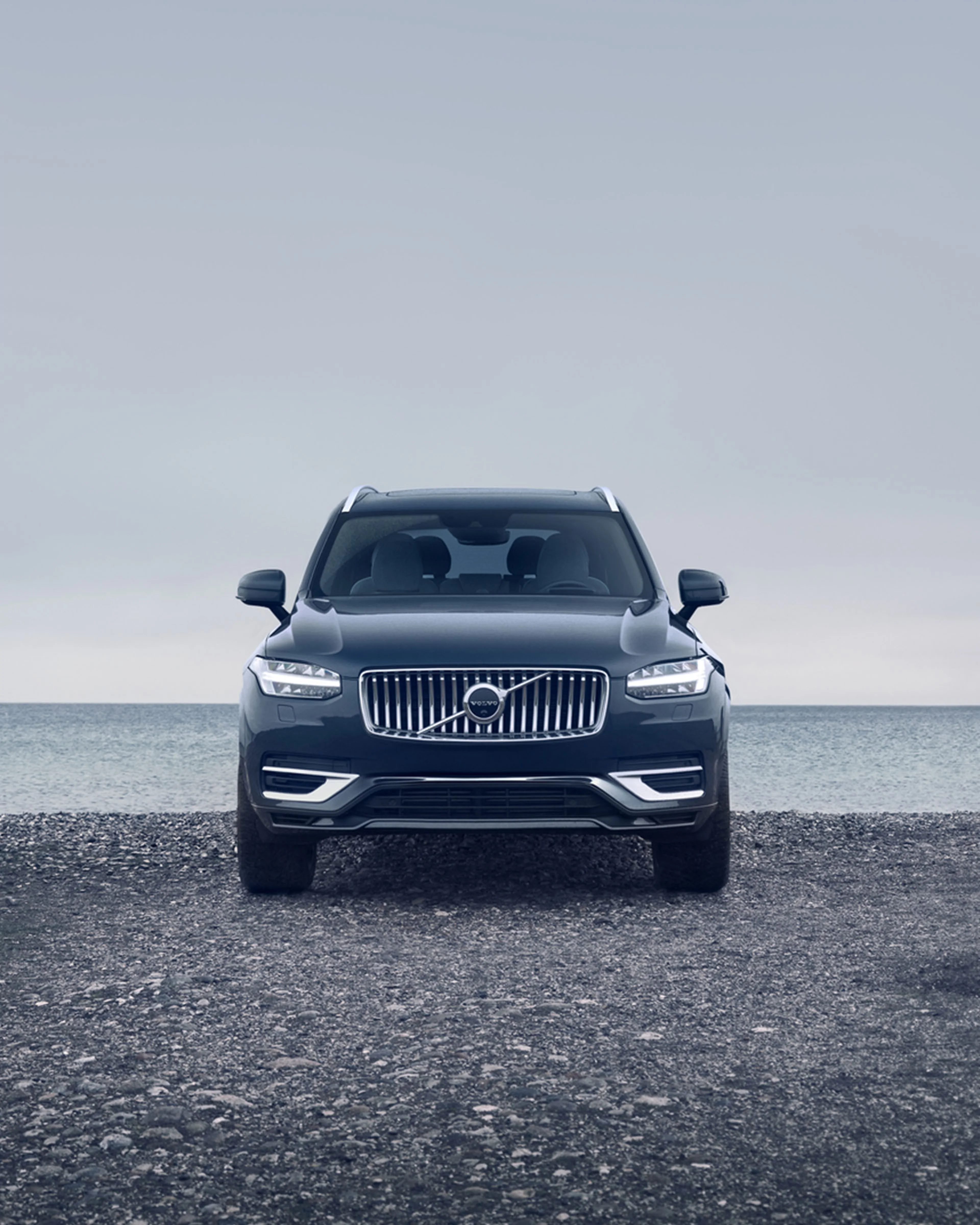 The front profile of a Navy XC90 on a pebble beach with a calm sea behind it