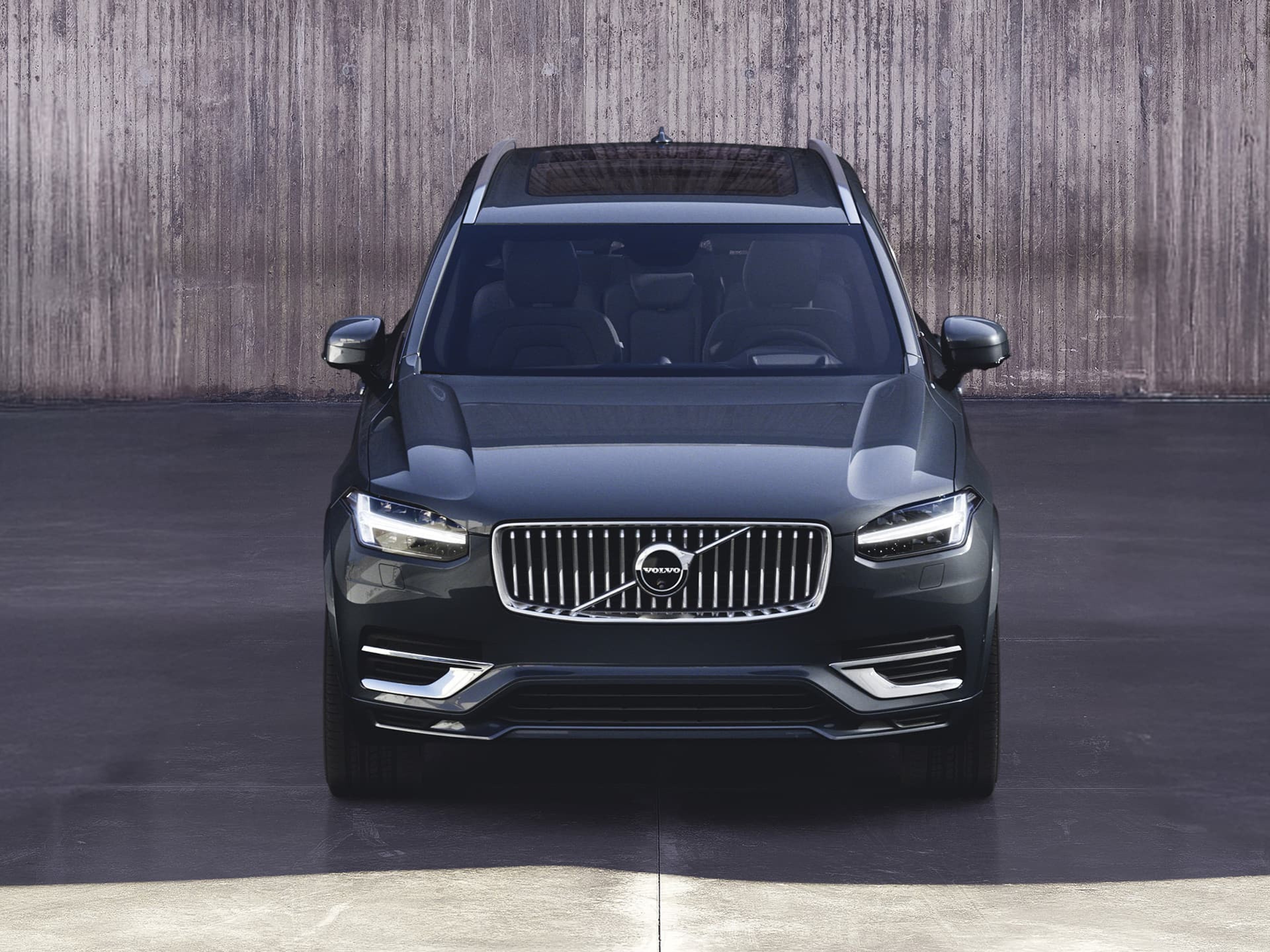 Vooraanzicht van een donkergrijze Volvo XC90 SUV, geparkeerd voor een betonnen muur.