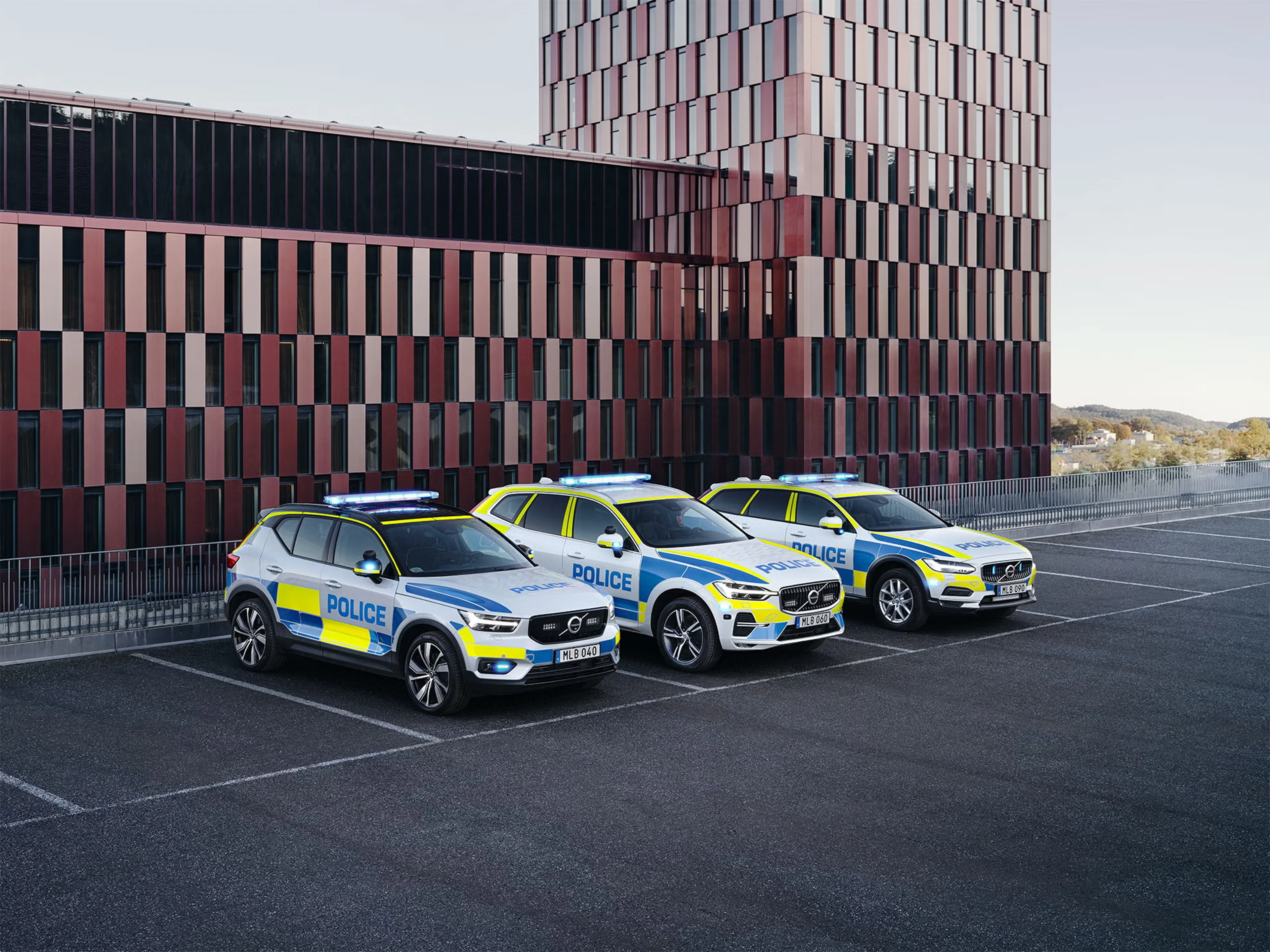 Tre Volvo politibiler i SUV-format med blå og gule markeringer holder parkeret foran en moderne bygning med lodrette stribede paneler og udstråler en effektiv og sikker stemning.