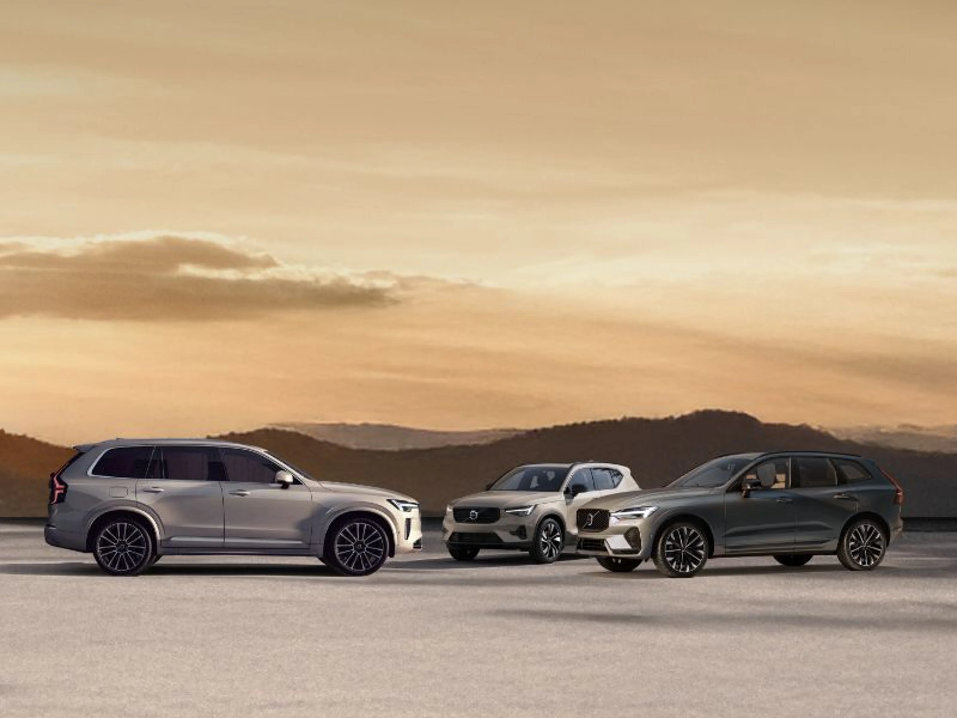 Tri Volvo SUV vozila parkirana na odprtem prostoru ob sončnem zahodu, z gorami v ozadju.