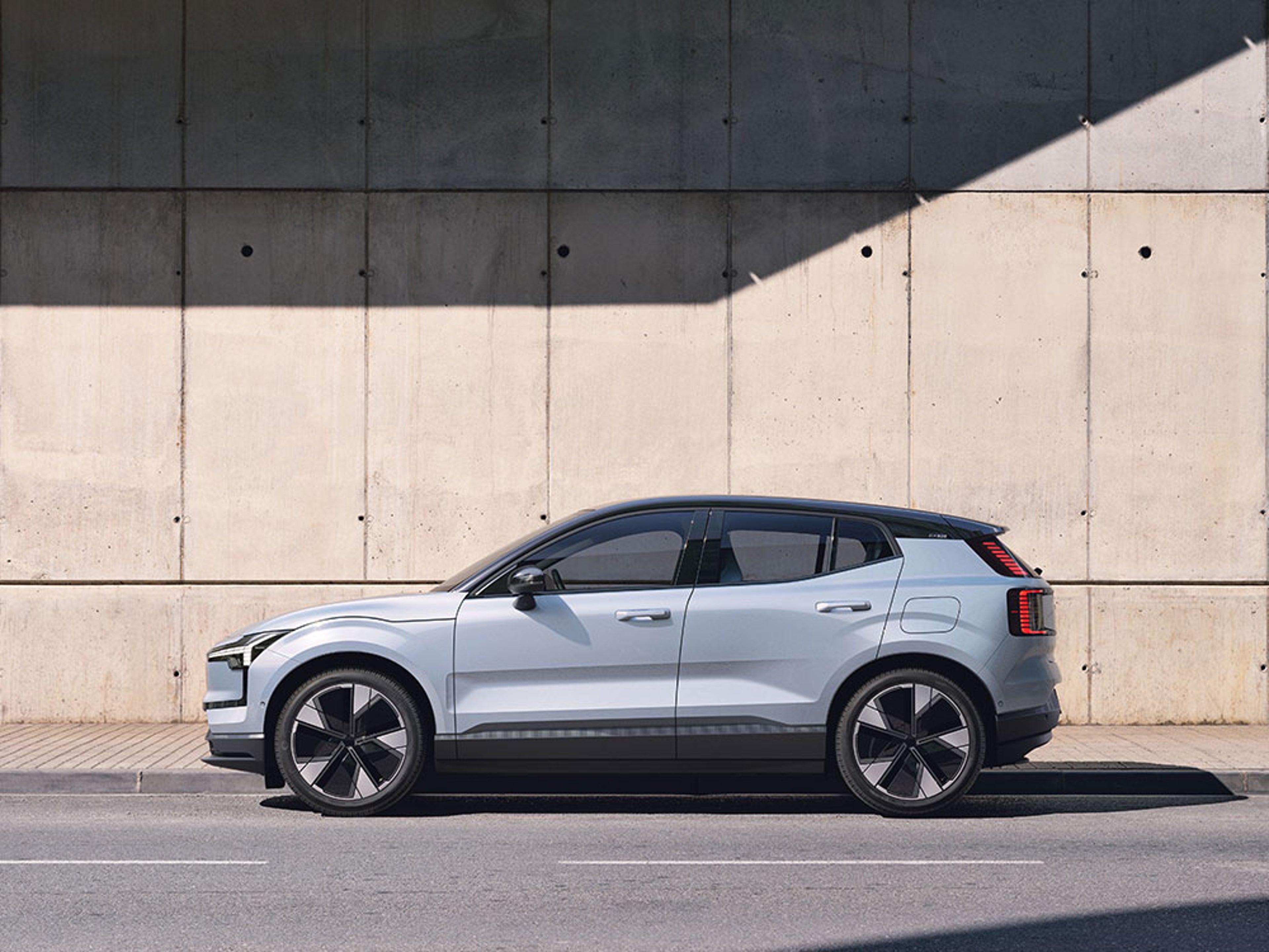 Zijaanzicht van een lichtblauwe Volvo EX40 elektrische SUV geparkeerd langs een betonnen muur.