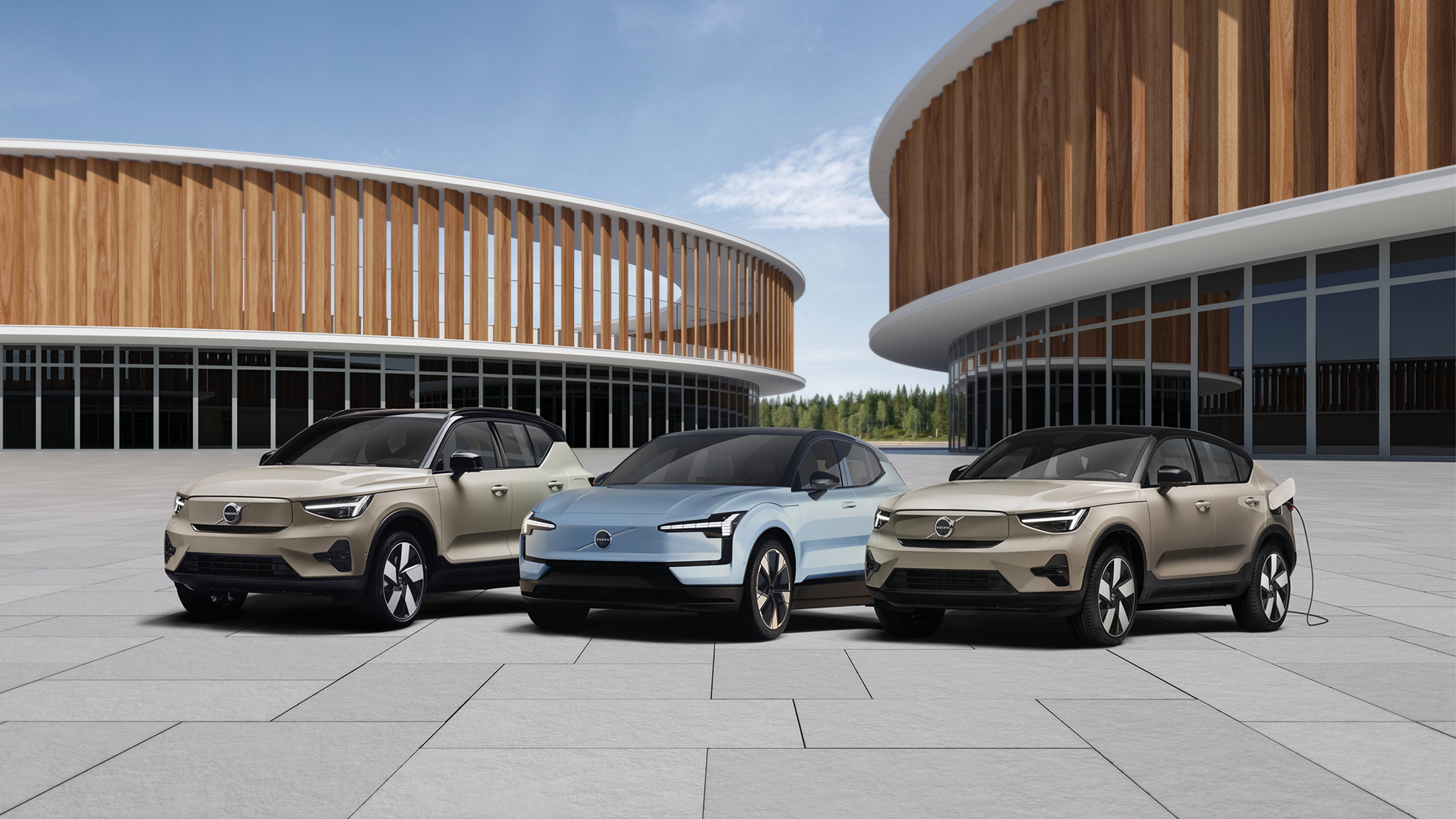 Trois SUV électriques Volvo garés devant un bâtiment moderne en bois et verre, sous un ciel dégagé.
