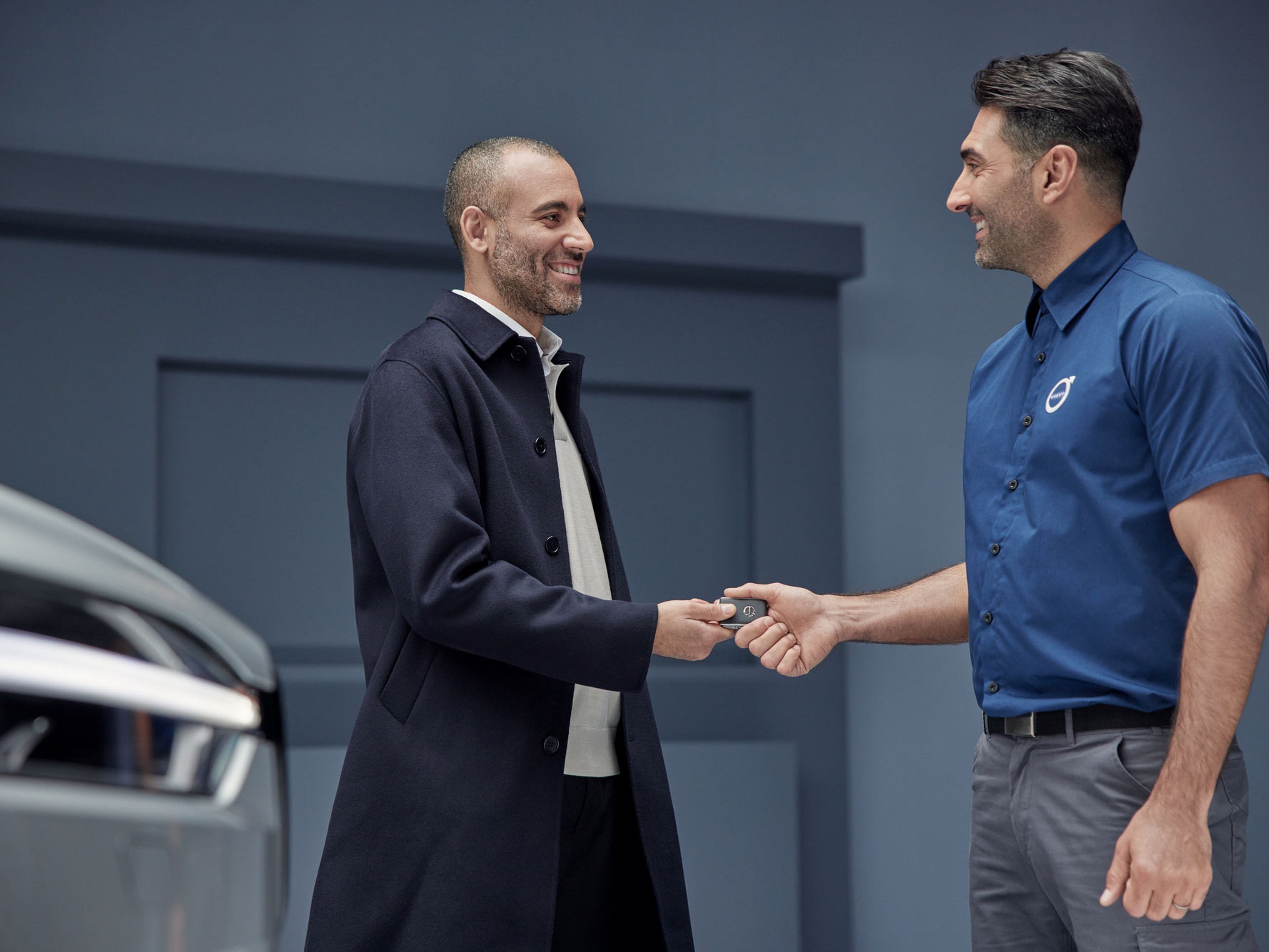 To mænd smiler og udveksler bilnøgler – den ene iført en mørk frakke, den anden en blå skjorte med Volvos logo. De står foran en sølvfarvet Volvo bil og udstråler en venlig stemning.
