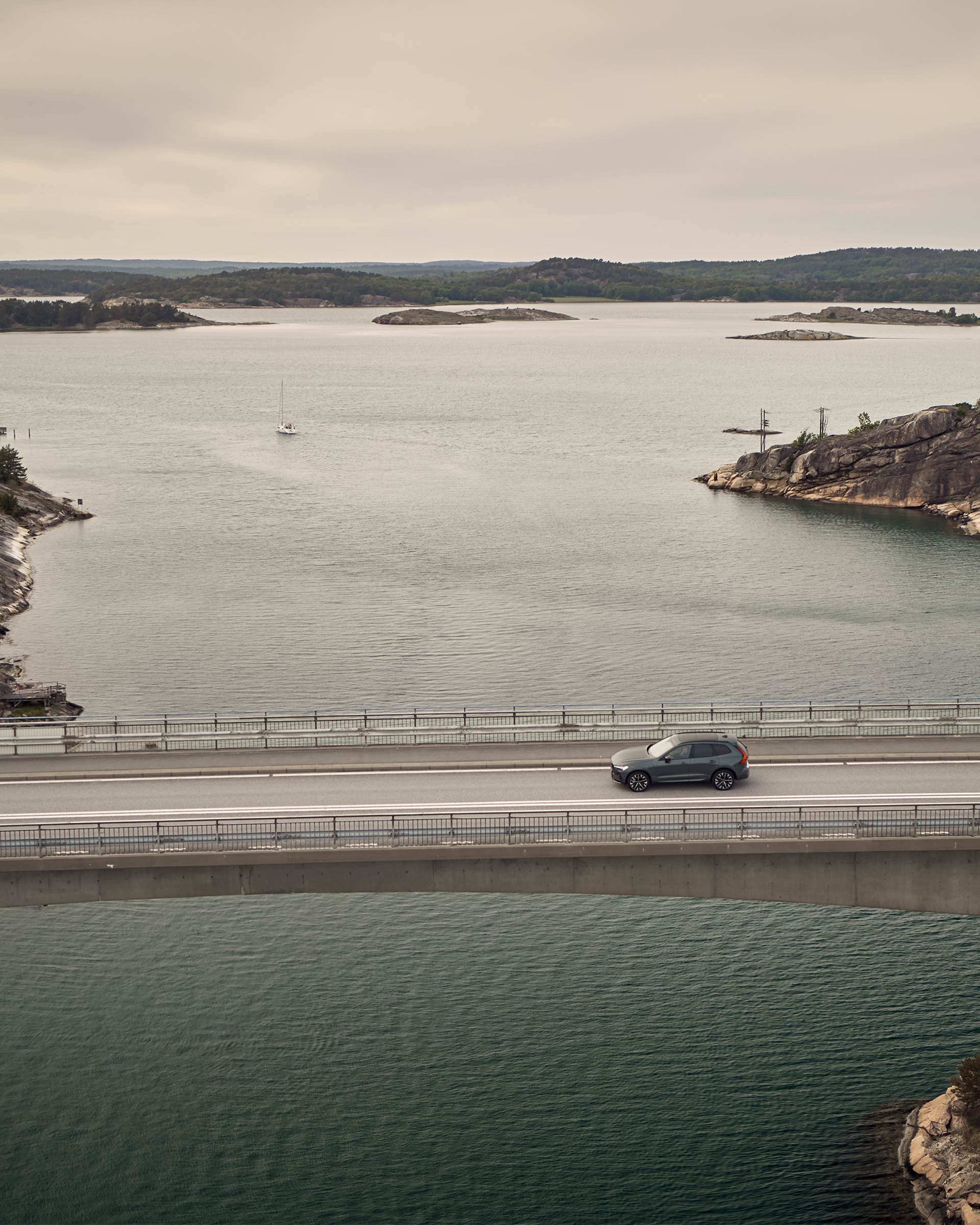 Forest lake -värinen Volvo XC60 lataushybridikatumaasturi ajaa vesistön ylittävällä sillalla. Taustalla saaristomaisema ja pois päin lipuva purjevene.