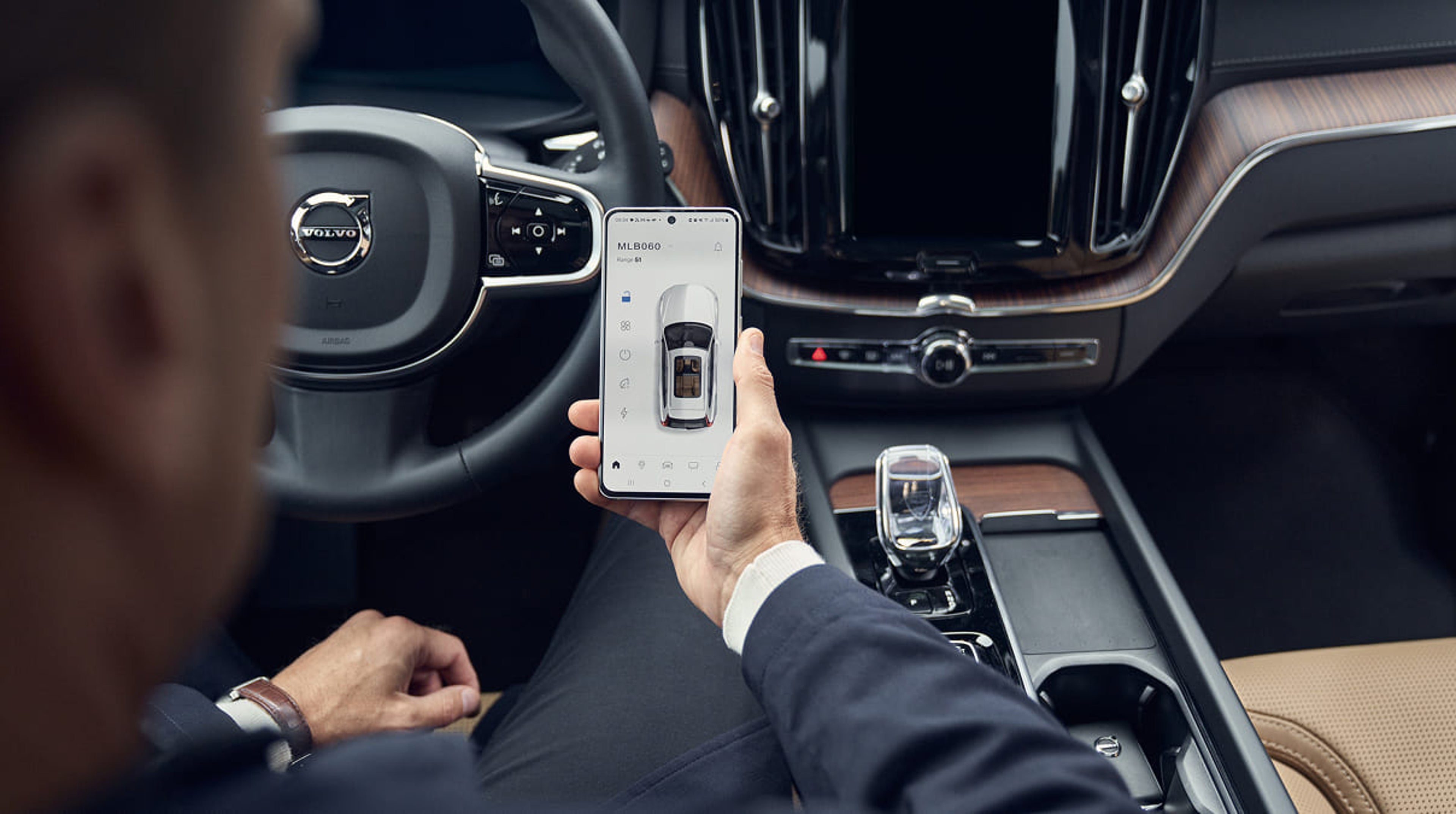 Hand holding a phone showing the Volvo Cars app. Steering wheel and dashboard in the background.