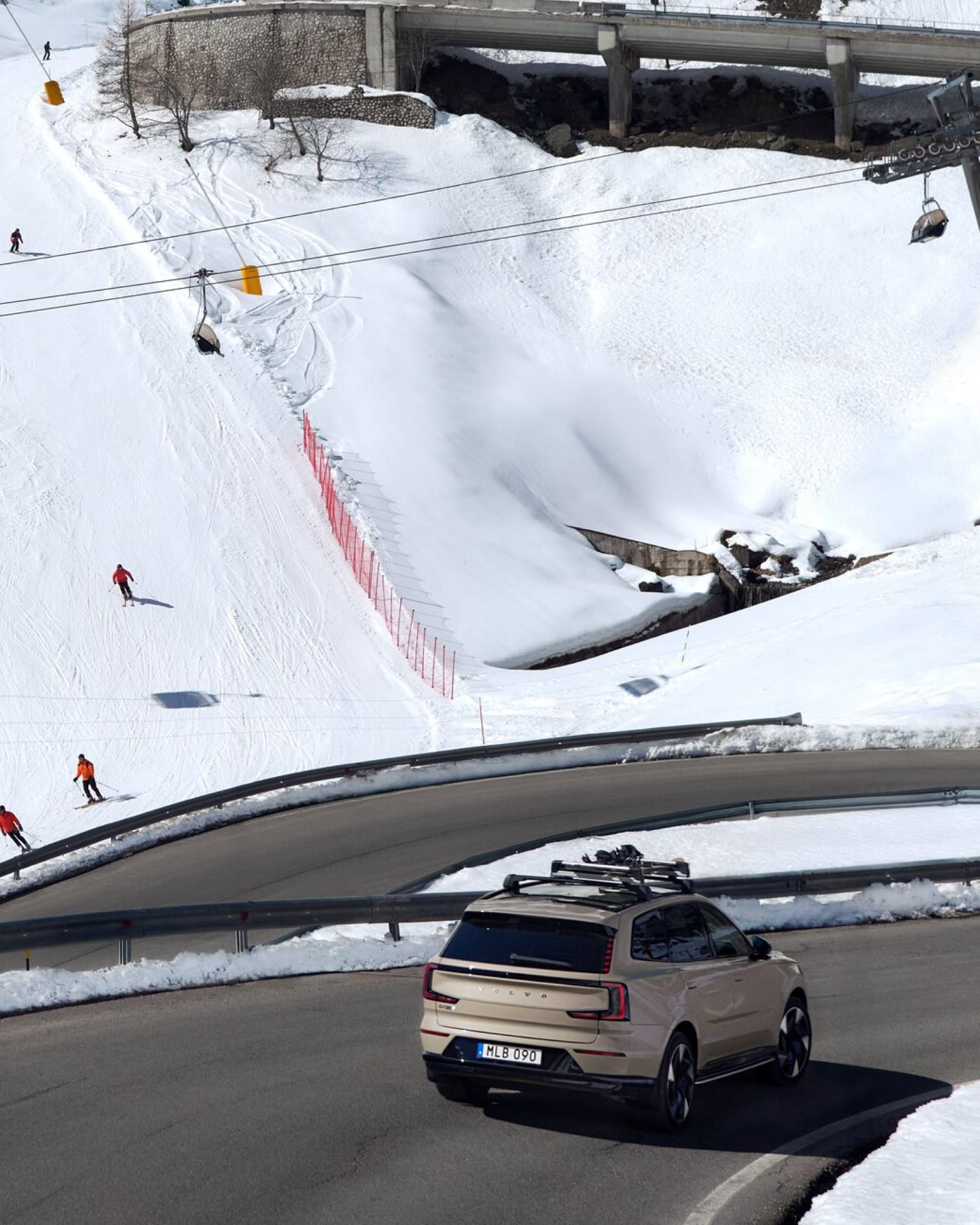 Een goudkleurige Volvo SUV met een skidrager op het dak rijdt op een kronkelende bergweg in een besneeuwd landschap, terwijl skiërs van de piste naast de weg afdalen.