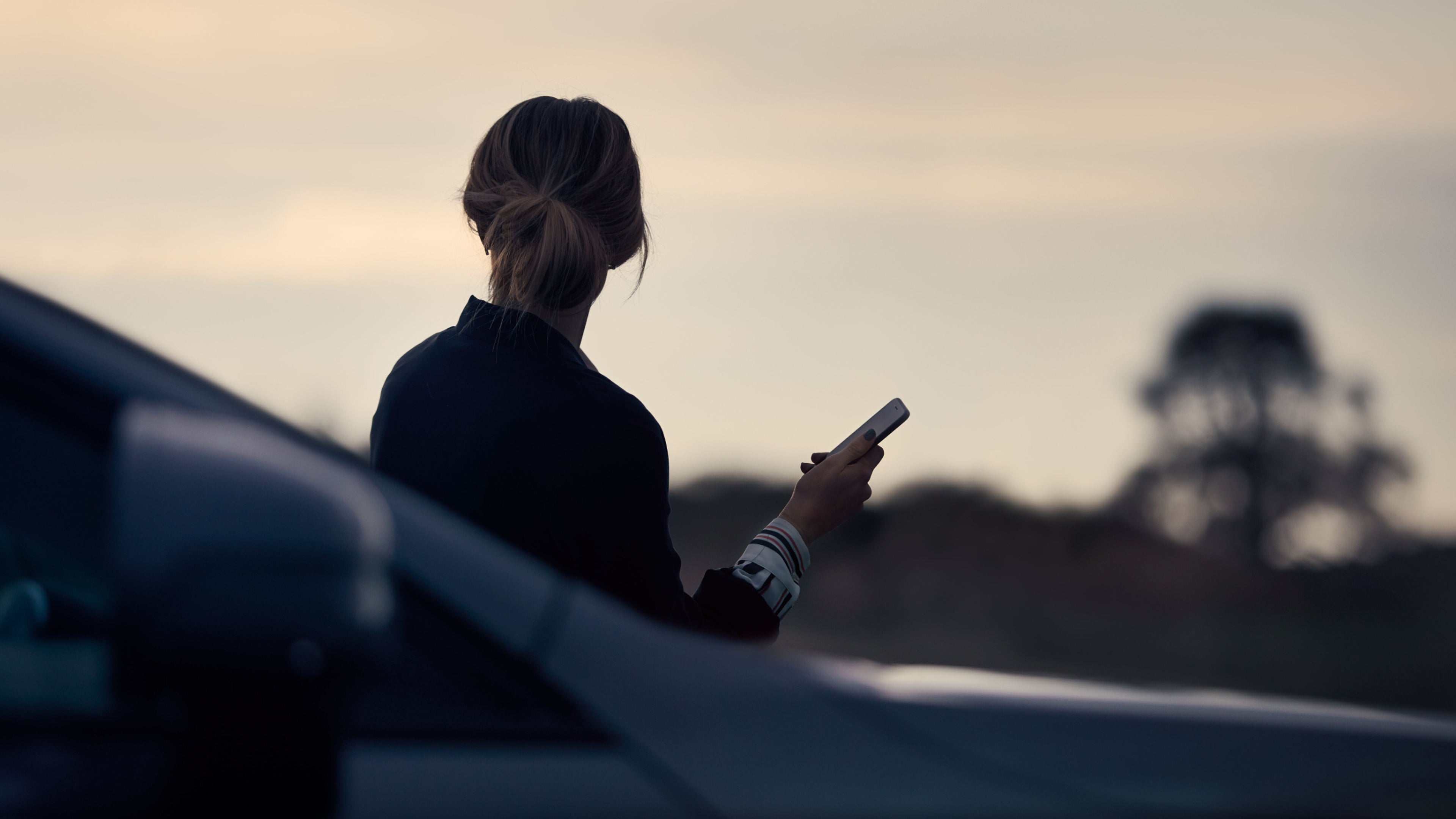 Eine Frau steht während der Abenddämmerung mit einem Handy an der Hand an einem Fahrzeug.