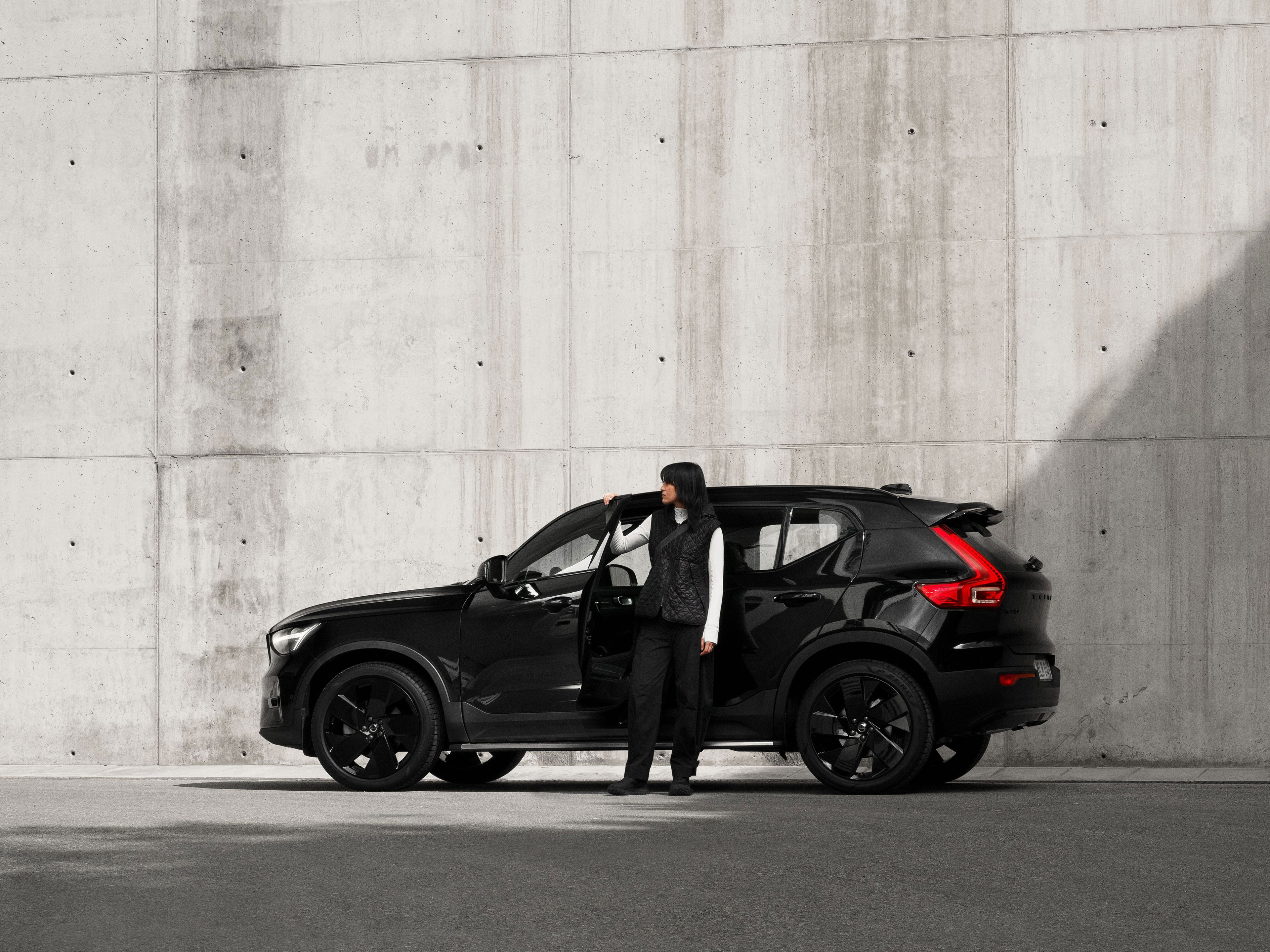 Eine Frau hat steht vor einem tiefschwarzen Volvo XC40 SUV, der vor einer grauen Steinmauer steht.