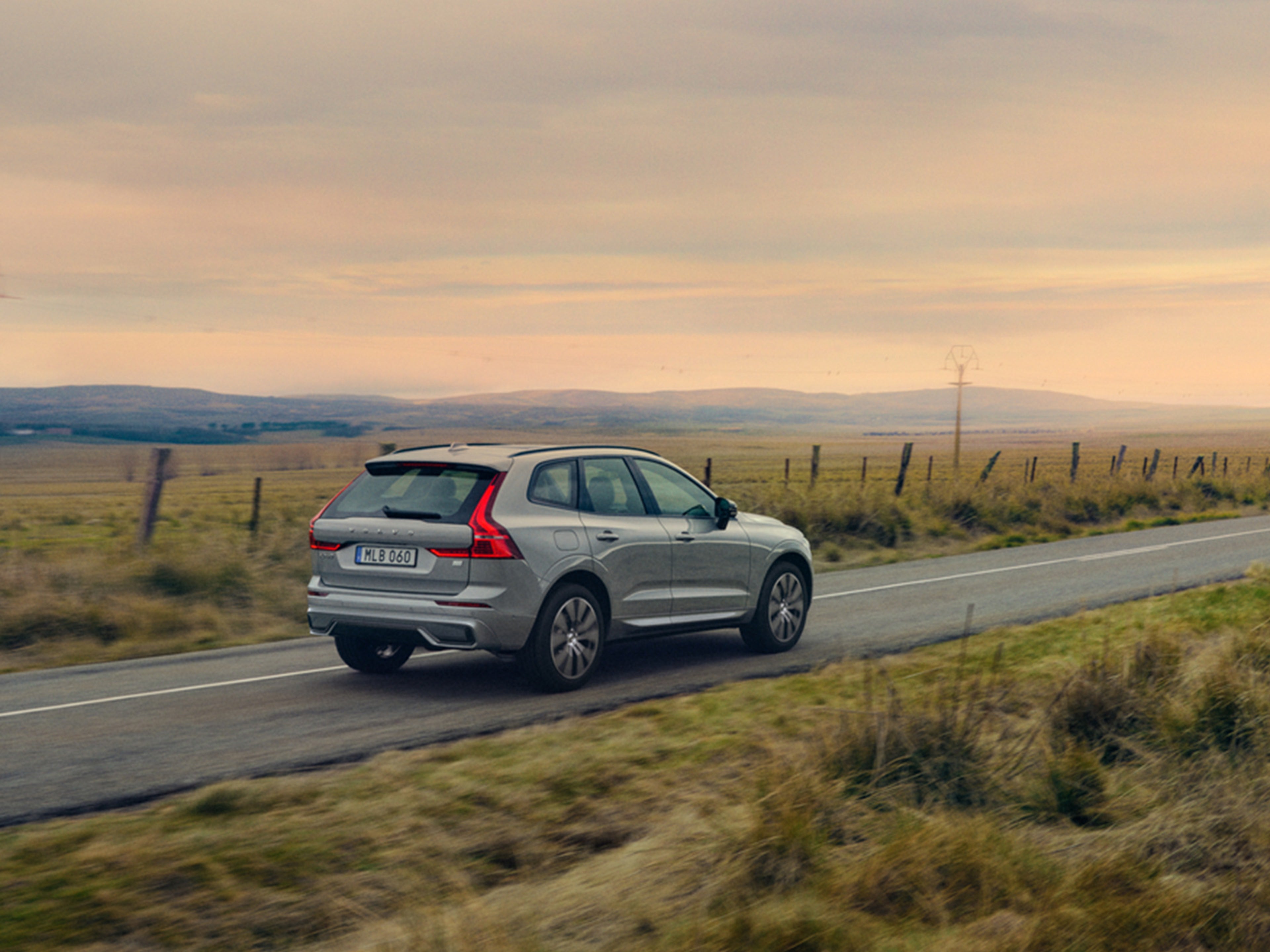 Silver Dawn -värinen Volvo XC60 lataushybridi ajaa peltojen välissä maantiellä auringonlaskussa.