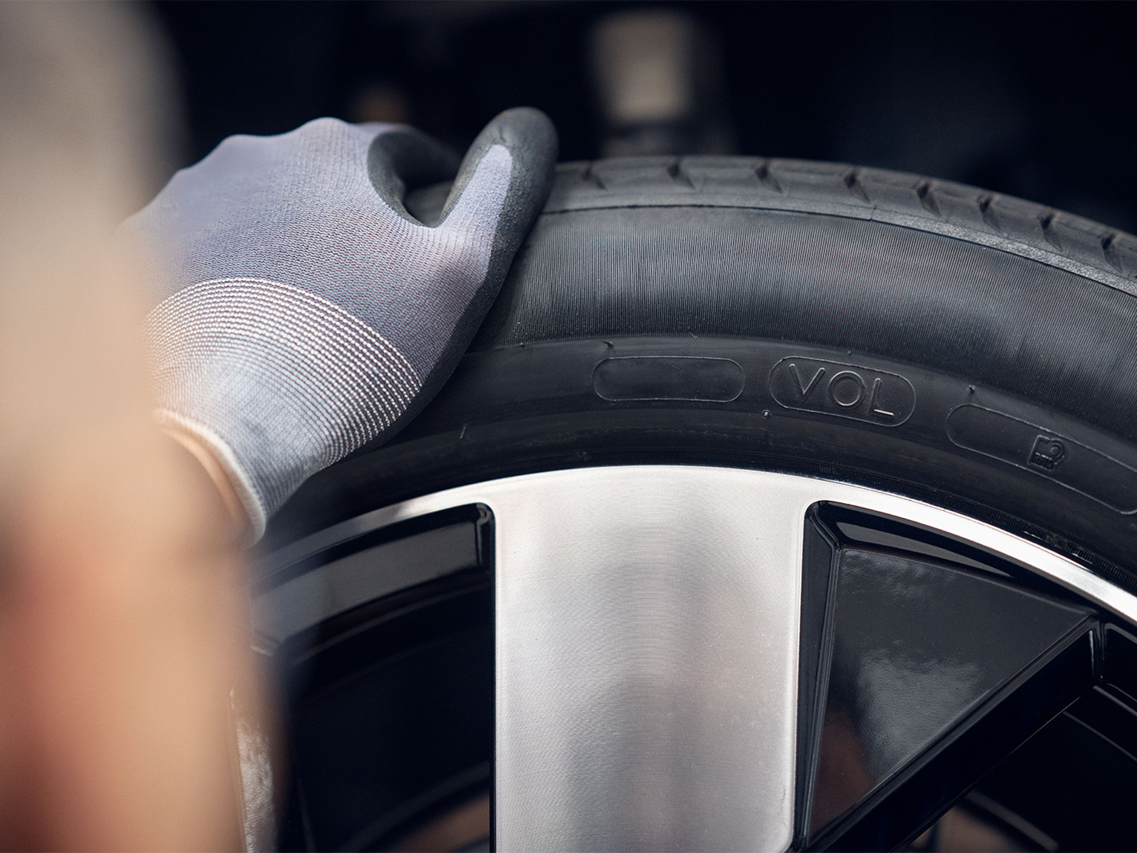 Close-up de uma mão enluvada a inspecionar um pneu de carro, com foco nos detalhes do piso e do aro, destacando a manutenção automóvel da Volvo.