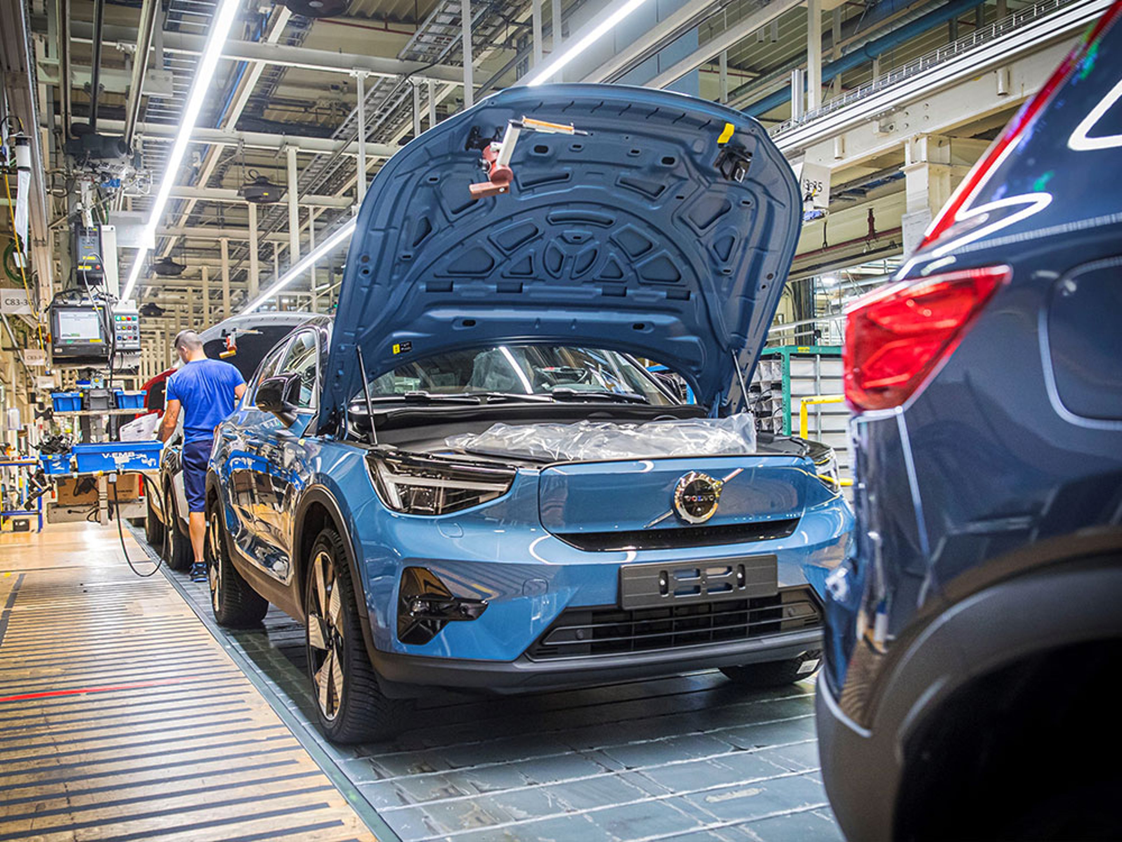Een blauwe Volvo EX40 met open motorkap op een productielijn in een autofabriek. Een arbeider staat bij de auto.