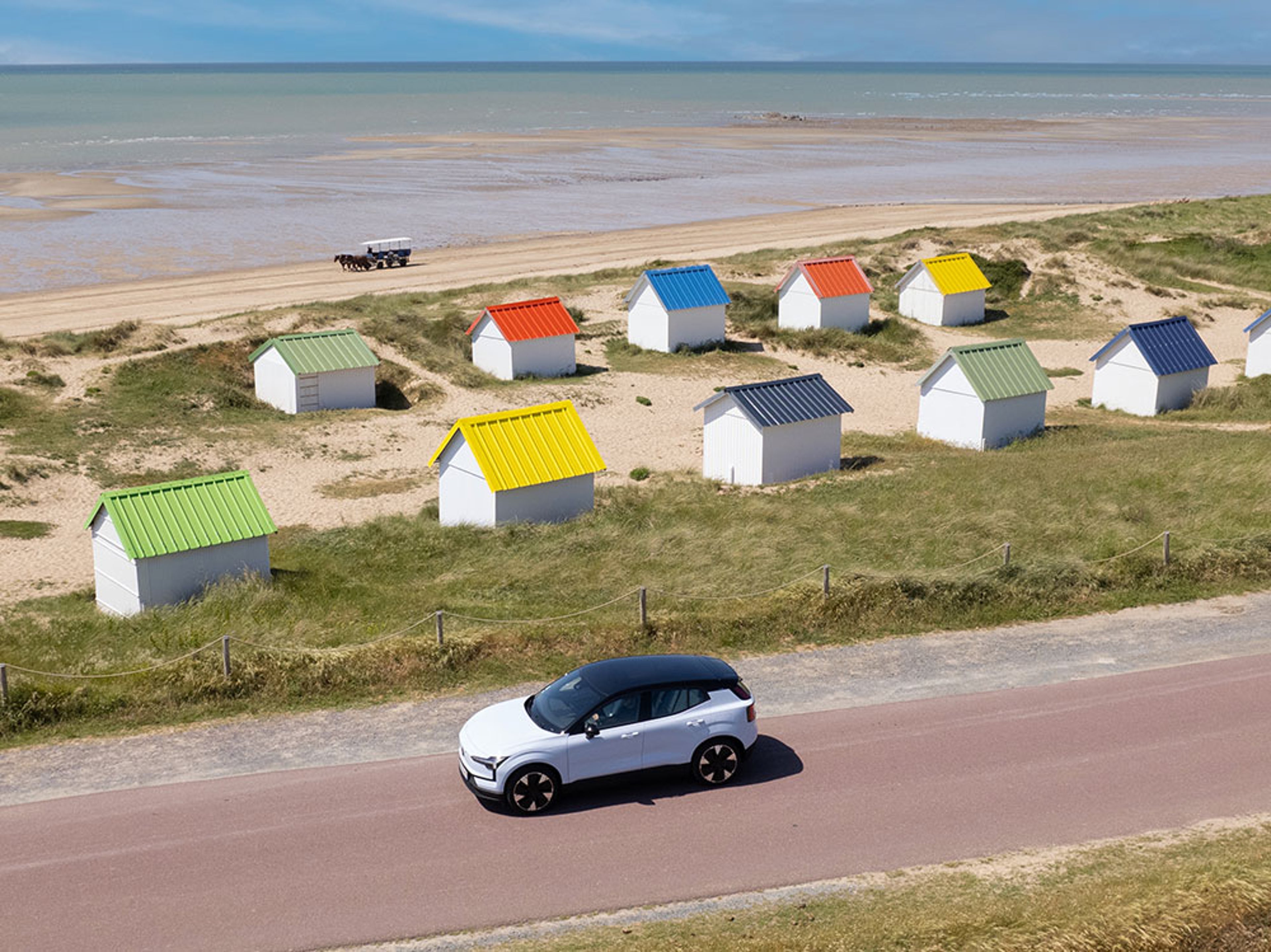 Vue aérienne de cabanes de plage colorées, route avec Volvo EX30, mer en arrière-plan