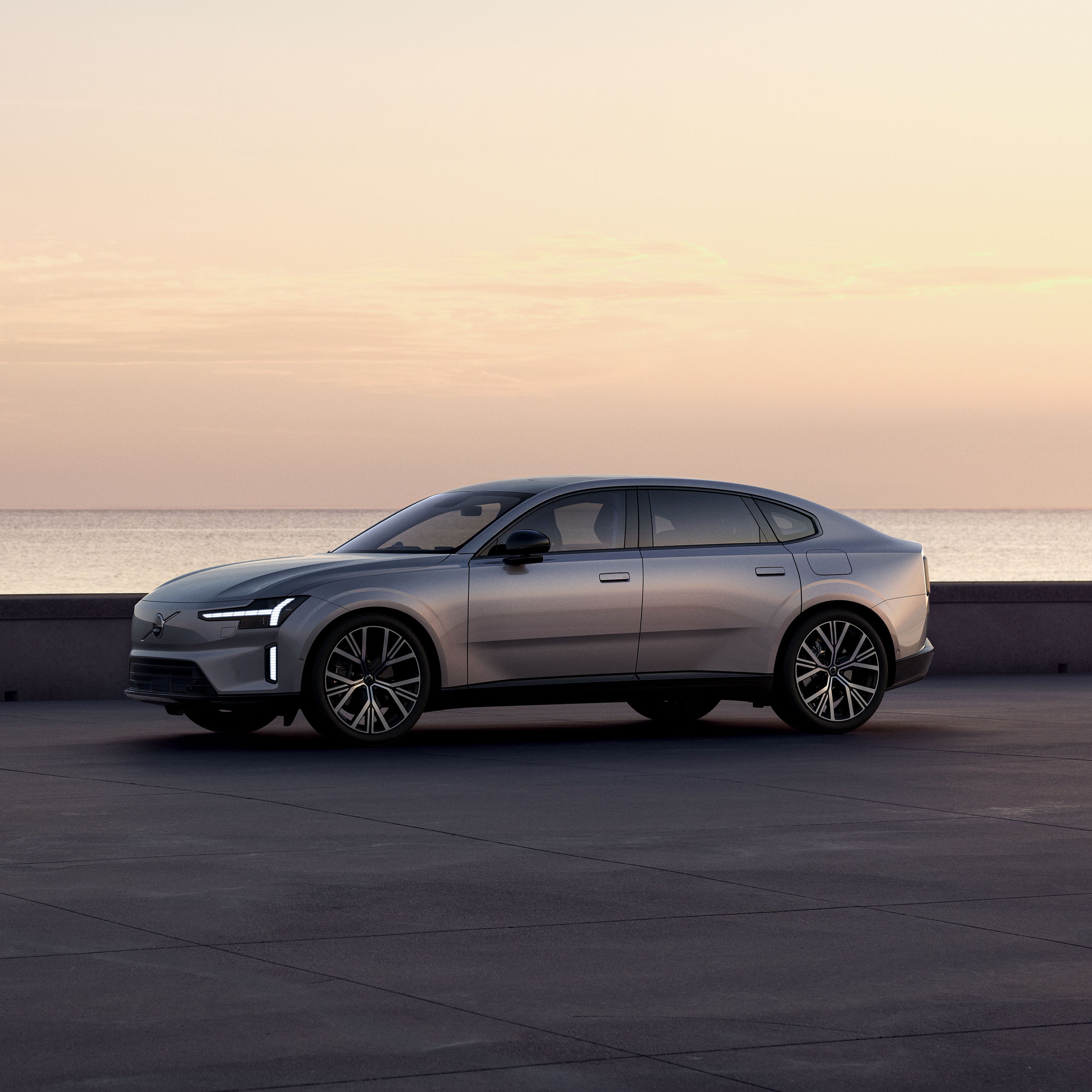 Aurora silver -värinen Volvo ES90 täyssähköinen sedanauto pysäköitynä rantatasanteelle auringonlaskussa.
