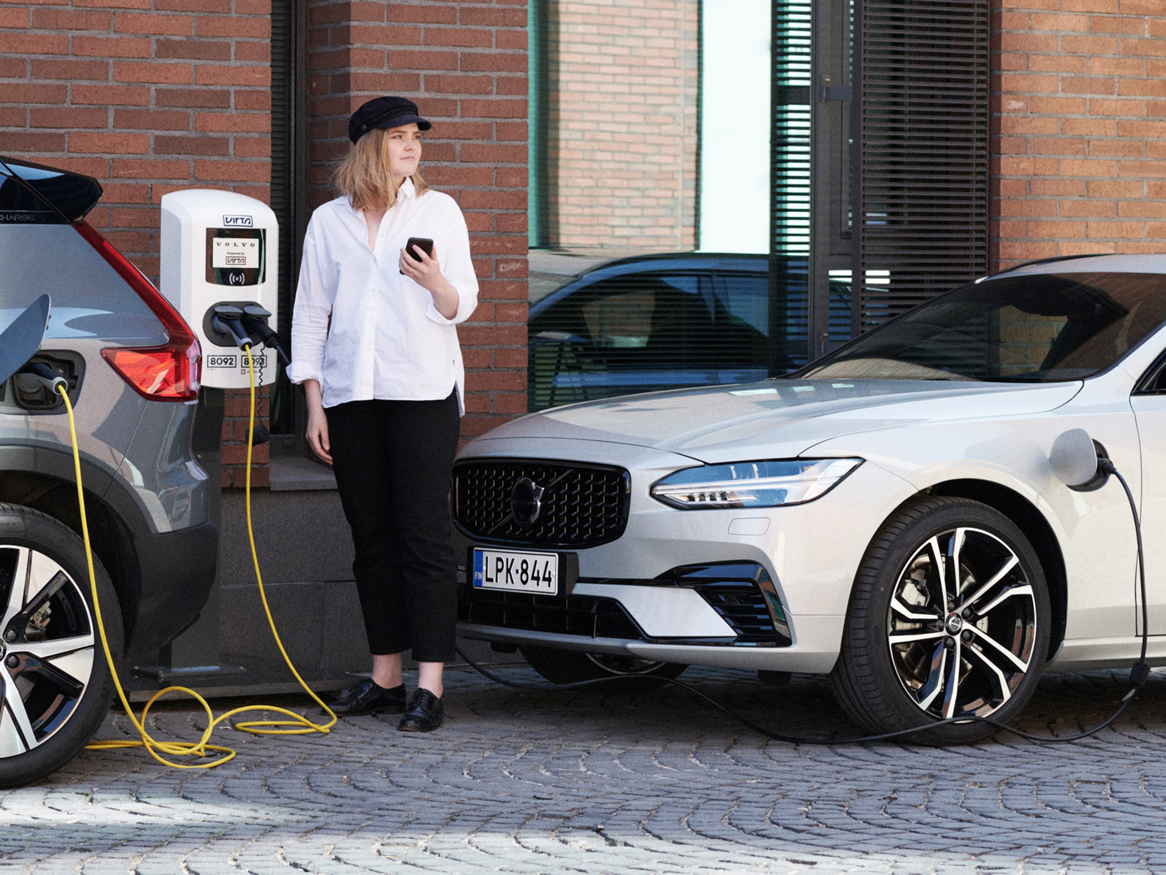 Nainen seisoo kahden Virta latausasemaan kytketyn Volvon välissä ja seuraa latauksen edistymistä puhelimestaan.