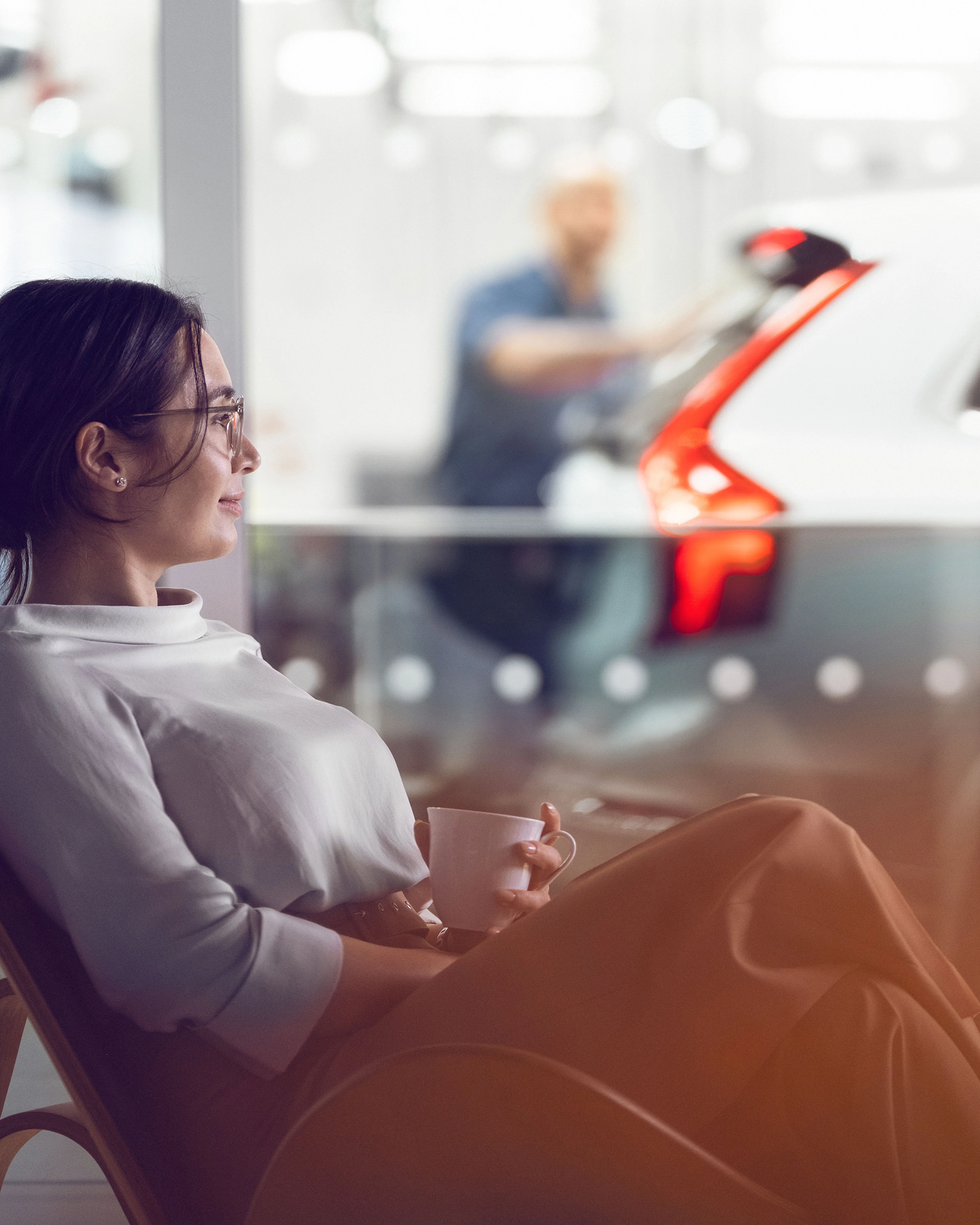 Eine Frau genießt einen Kaffee, sitzend im Wartebereich eines Showrooms vor einer Glasscheibe. Hinter der Glasscheibe steht ein neuer Volvo XC60.