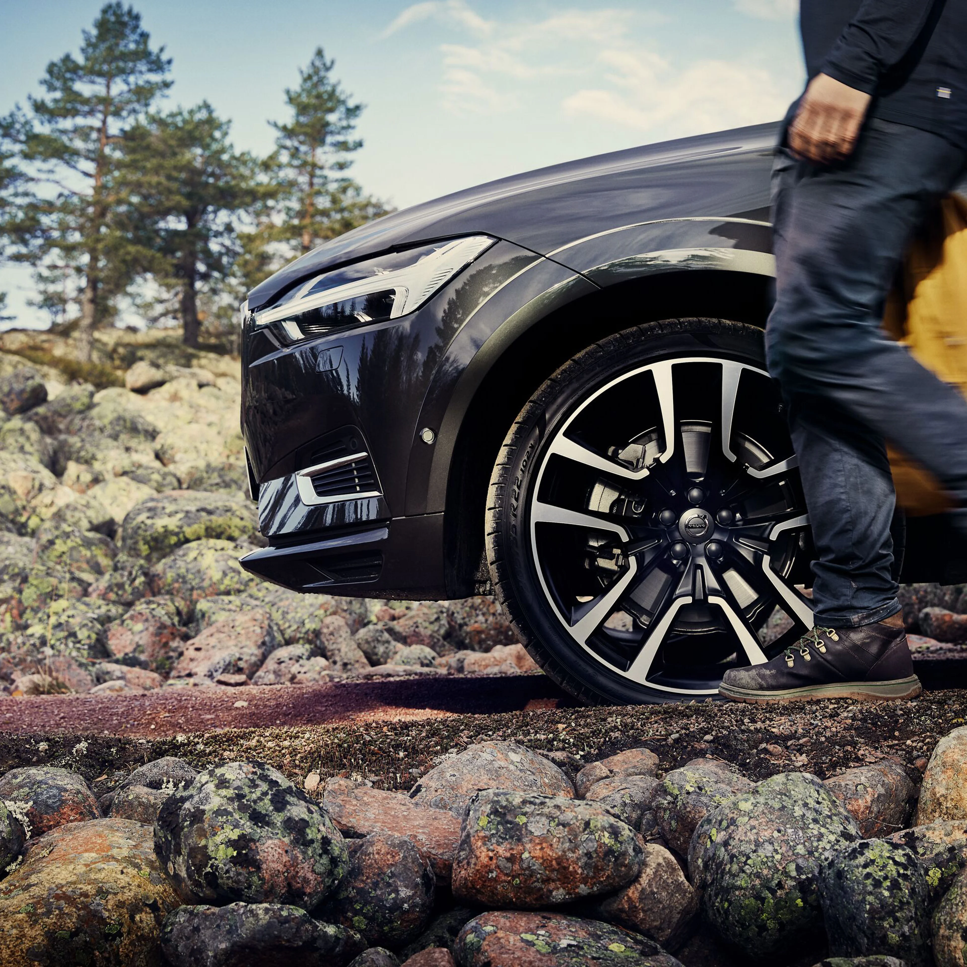 En person går forbi en sort Volvo, der er parkeret på klippefyldt terræn, med træer i baggrunden under en delvist skyet himmel.