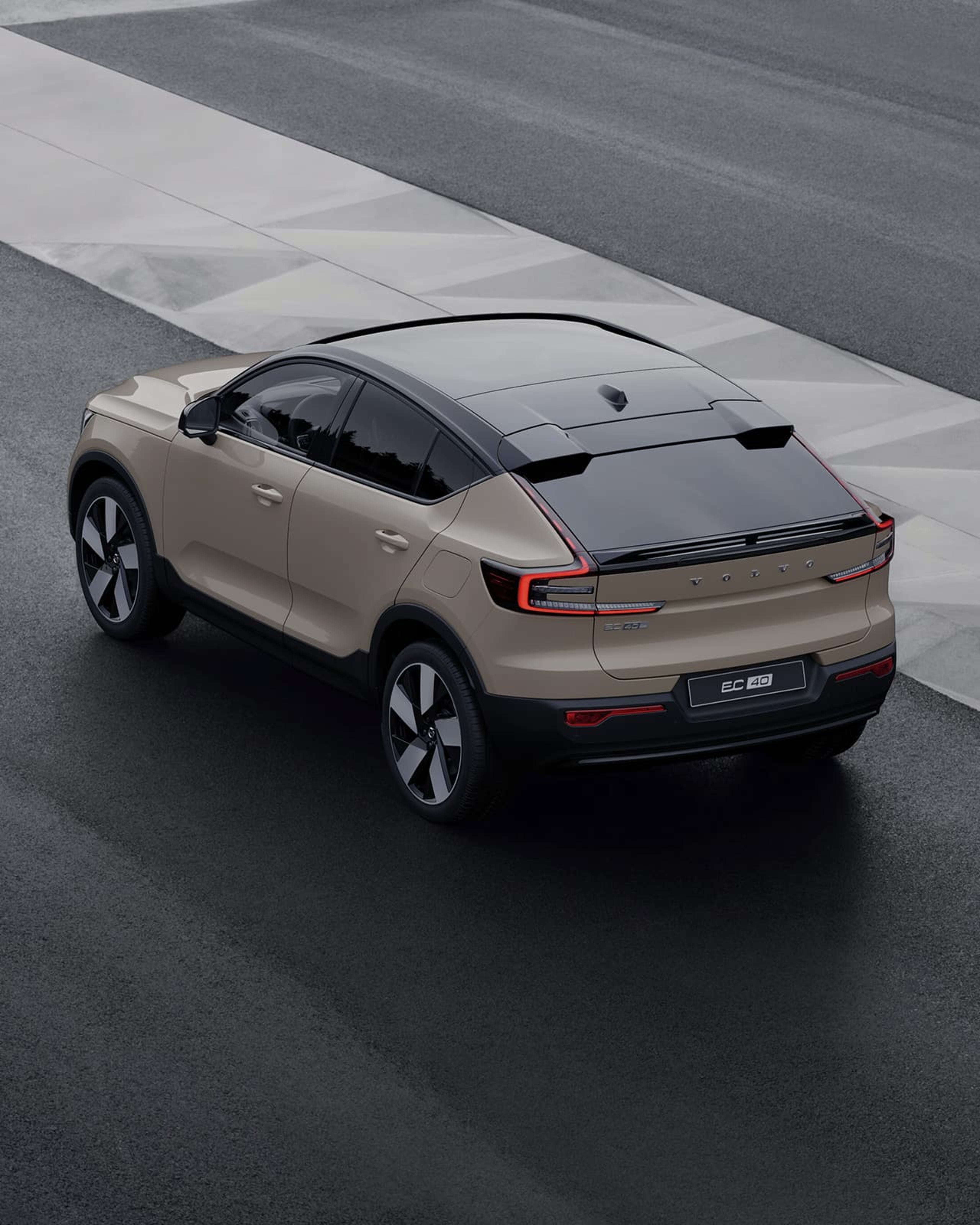 Vue arrière en plongée d'une Volvo EC40 de couleur beige sur une route asphaltée gris foncé et humide, avec des marquages géométriques gris clair.