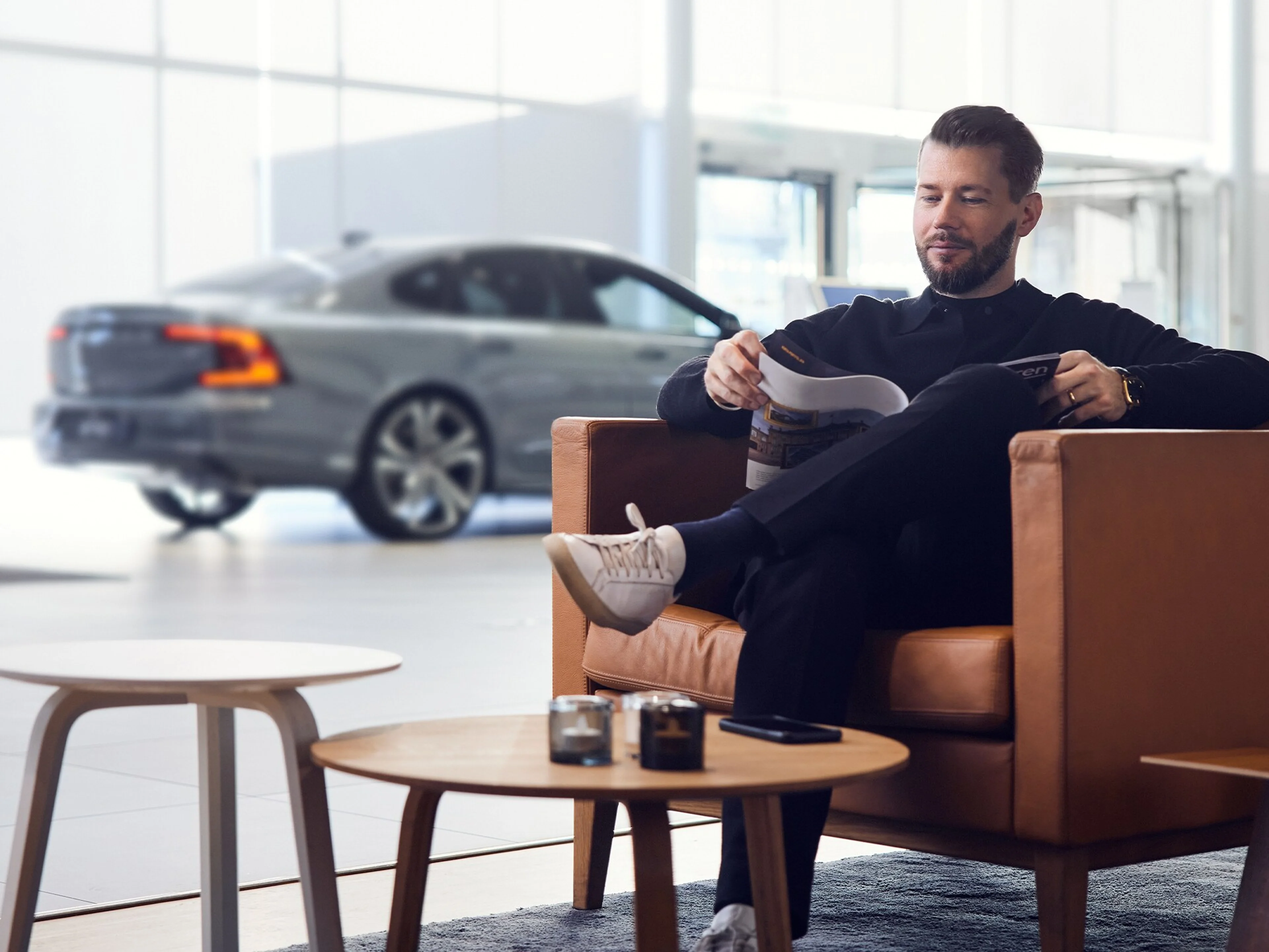 A person sits comfortably on a Scandinavian brown couch, reading a magazine with a Volvo visible through a large window behind them.