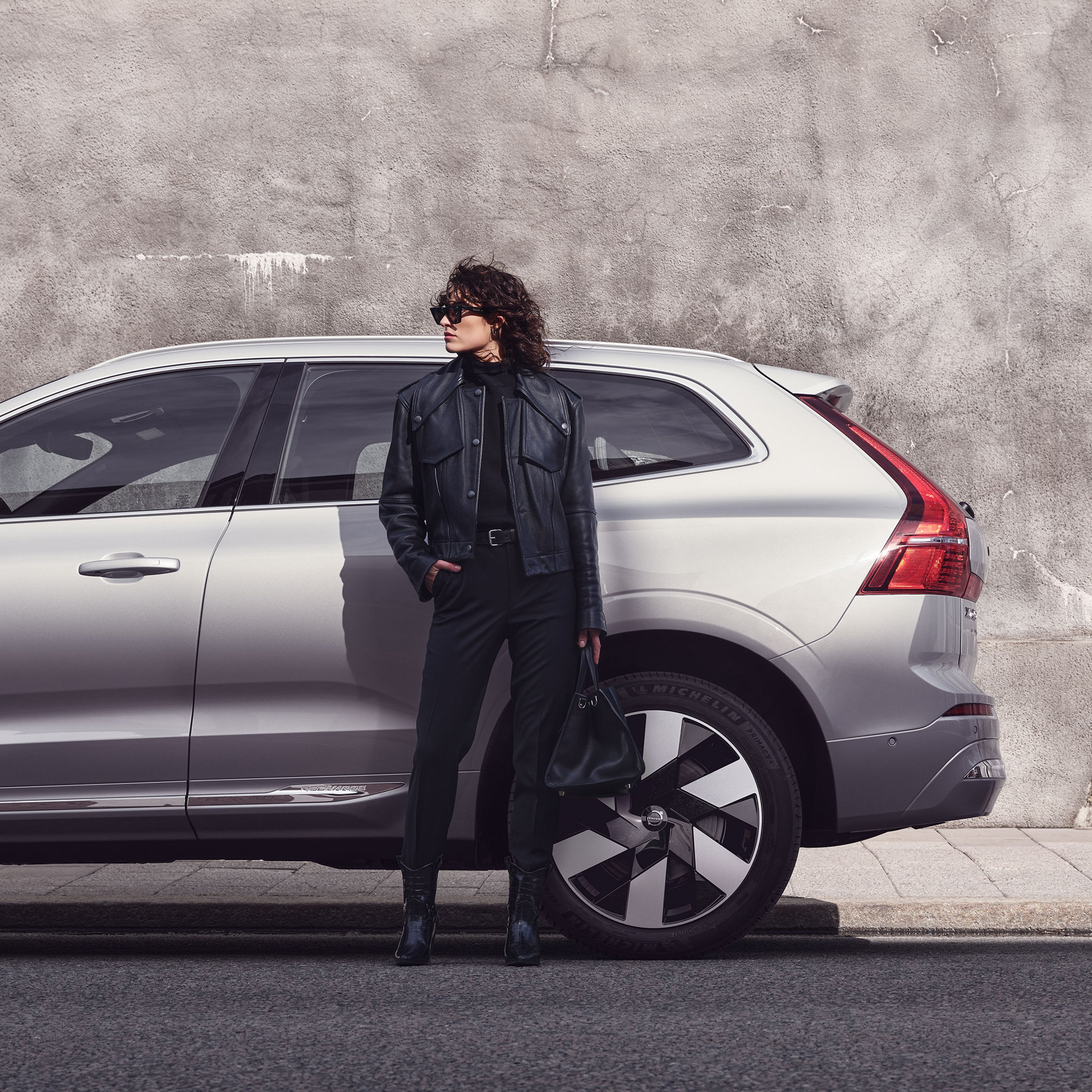 Mustaan asuun pukeutunut nainen seisoo käsi taskussa harmaan Volvo XC60 lataushybridiauton edessä ja katsoo sivulle.