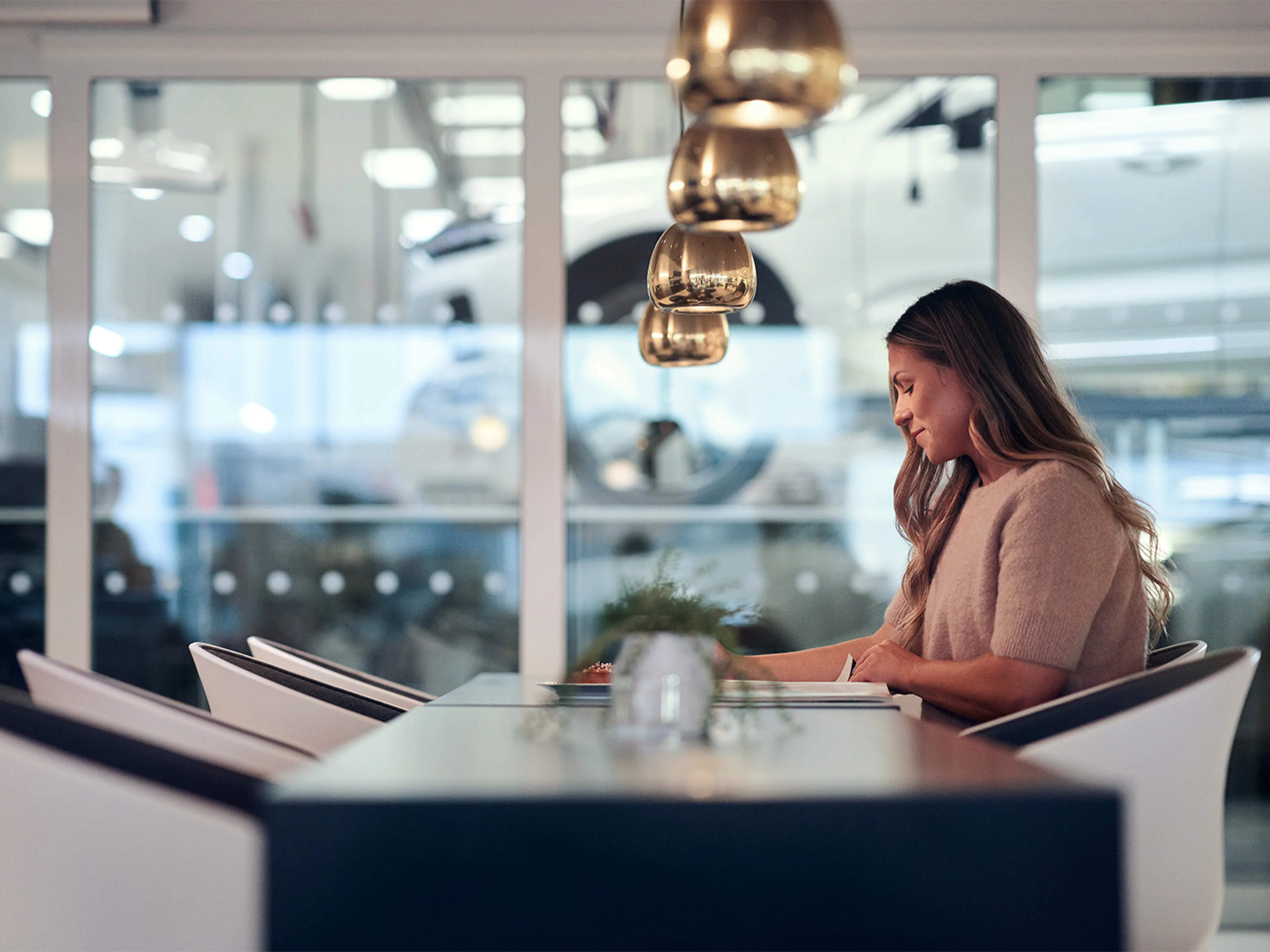 A woman sits at a modern table in a bright space, a Volvo showroom , with pendant lights overhead and a plant centerpiece.