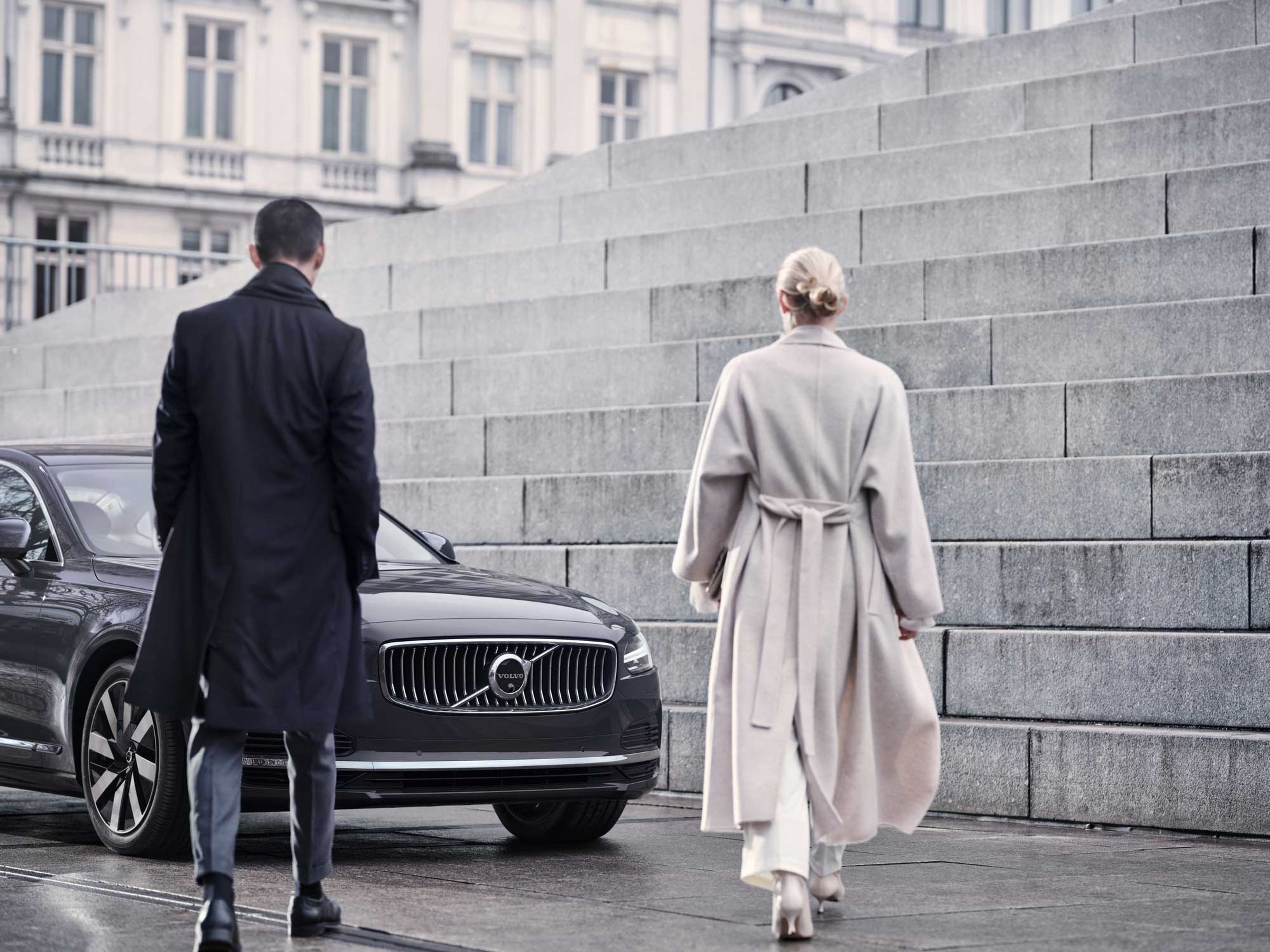 En mand og en kvinde går hen mod en mørkegrå Volvo parkeret ved store betonstrin, hvilket giver et professionelt og sofistikeret indtryk. Byens omgivelser antyder en forretningskontekst.