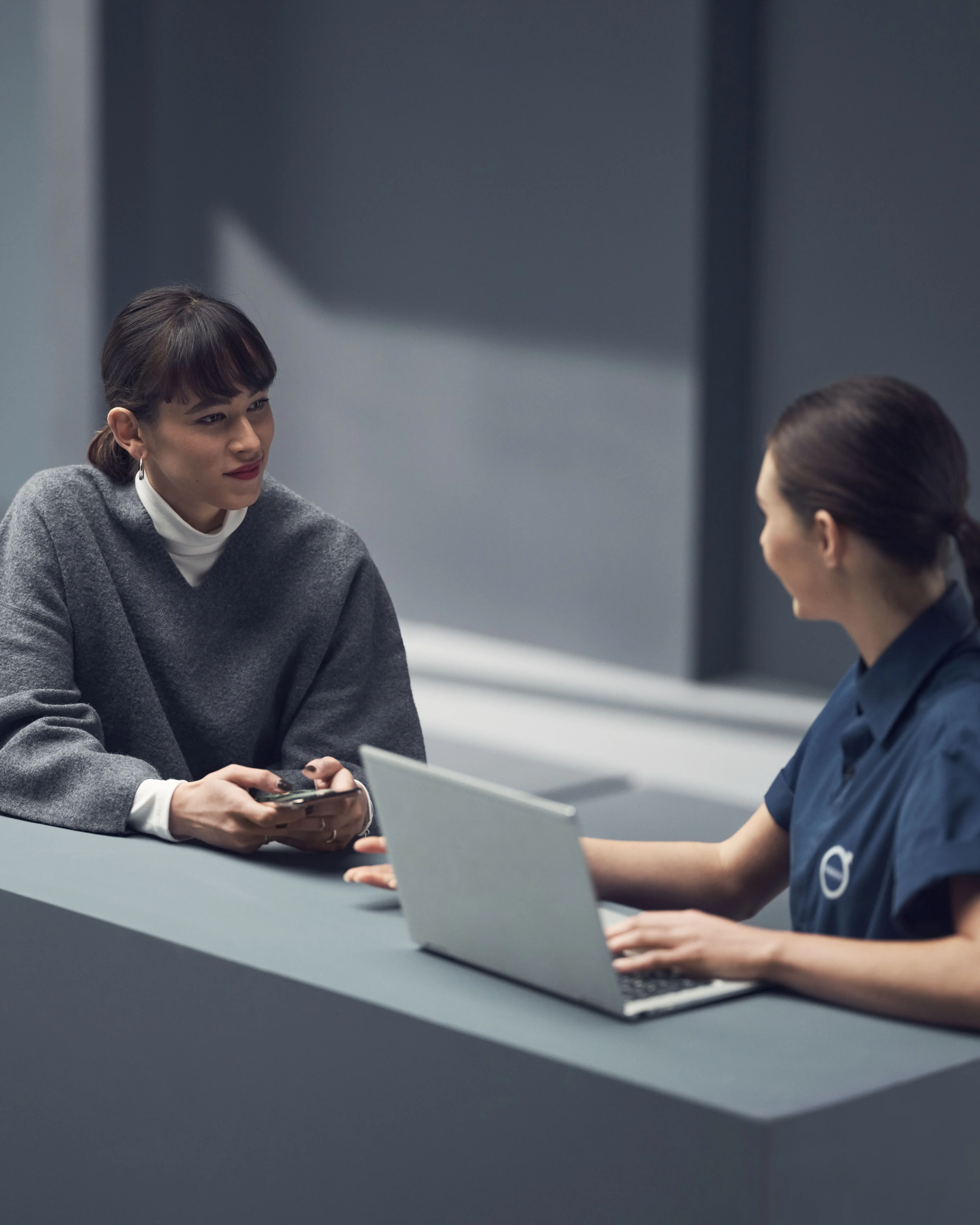 Zwei Frauen, eine mit einem Smartphone in der Hand, die andere mit einem Laptop vor sich, stehen sich gegenüber.