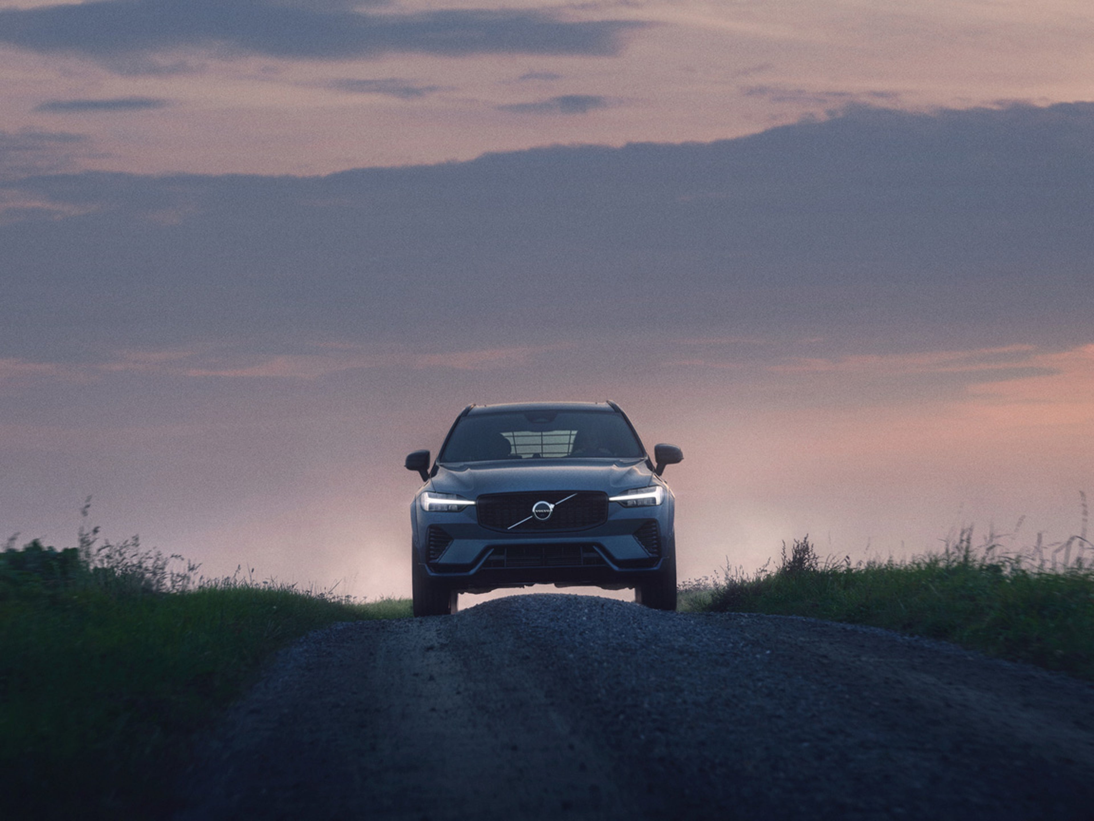 A Volvo XC60 SUV drives down a gravel road at dusk, surrounded by greenery under a colourful sky.