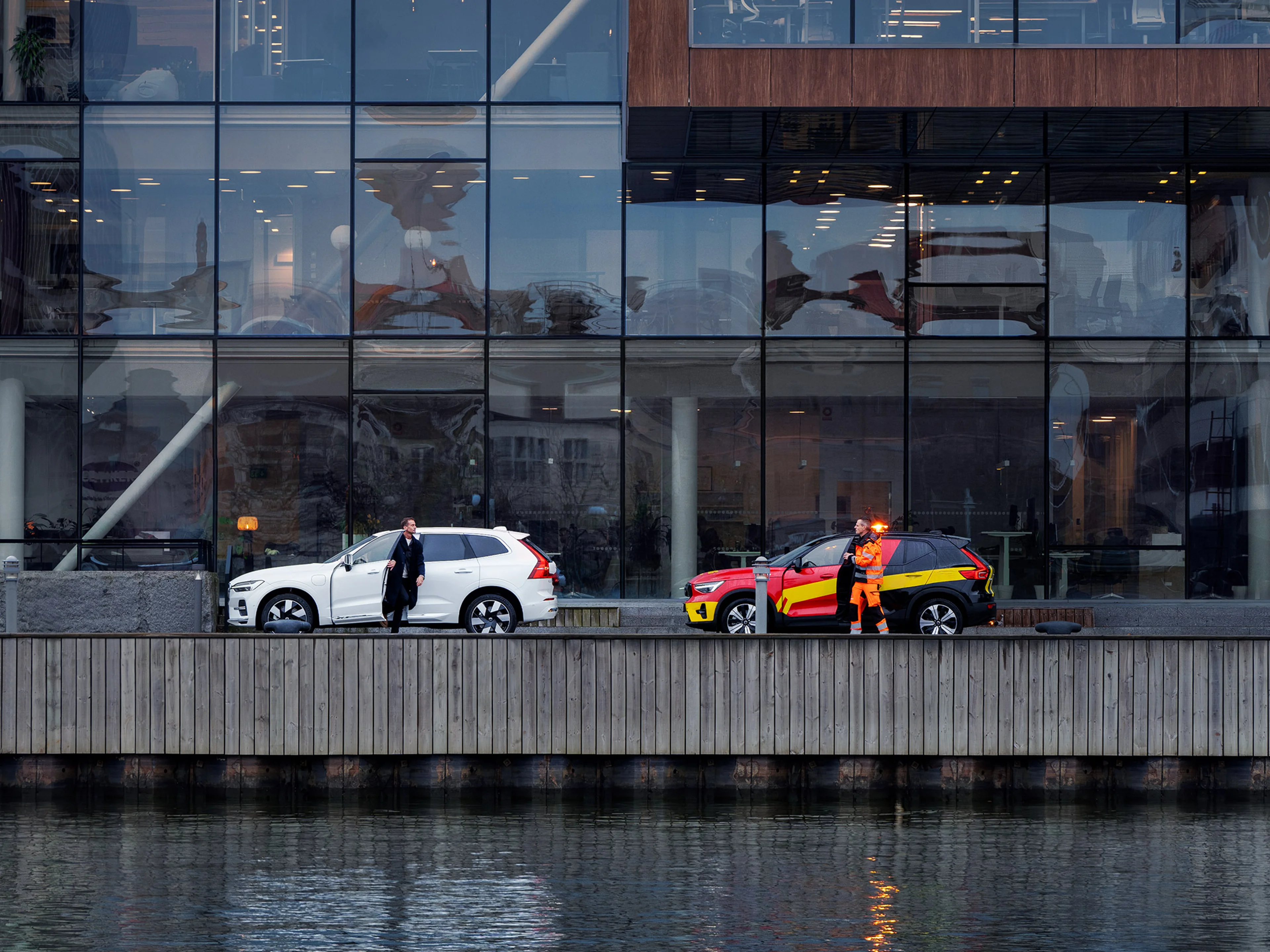 Valkoinen Volvo ja tiepalvelun auto pysäköitynä veden äärellä sijaitsevan toimistotalon edustalla.