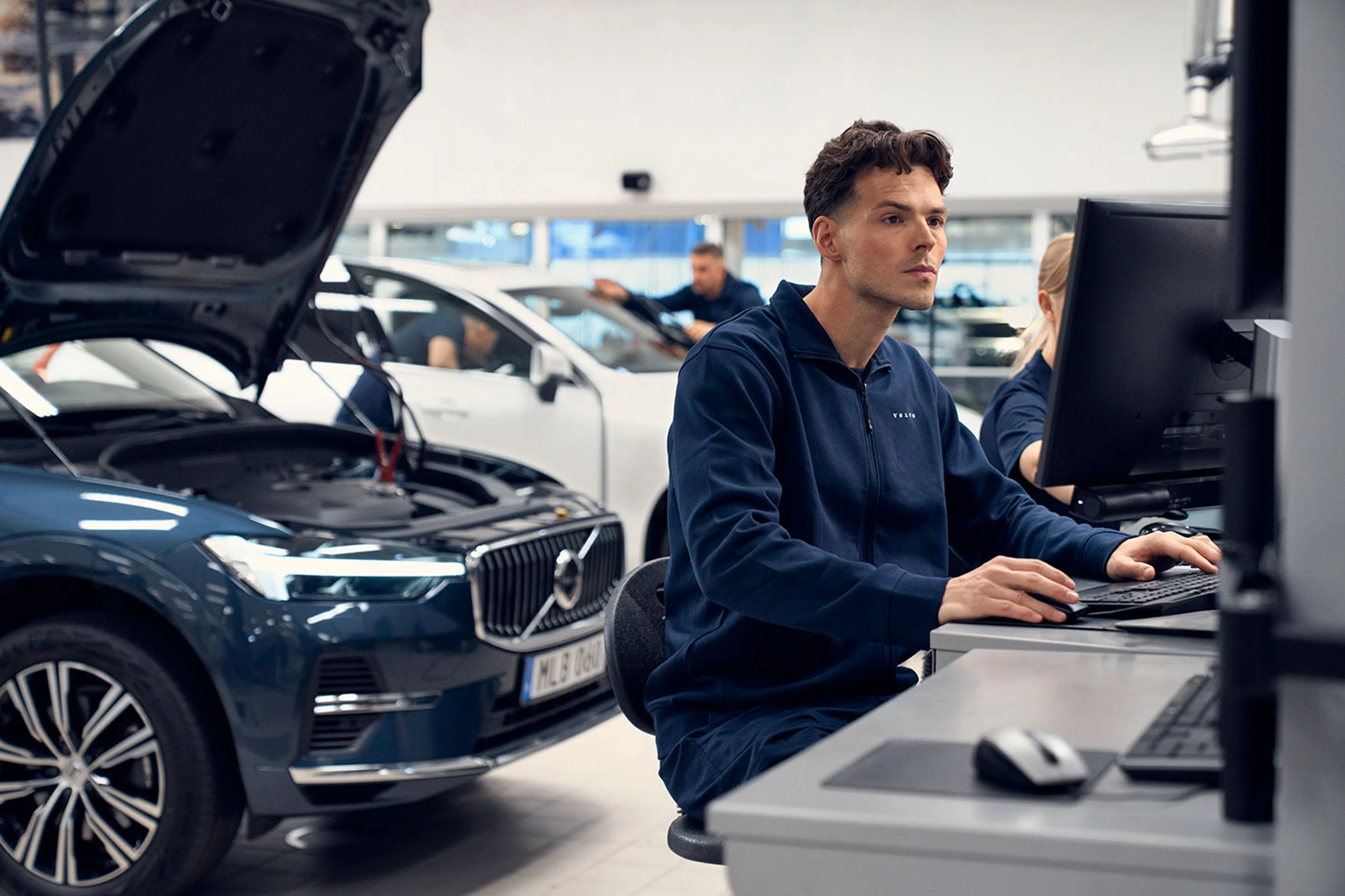 En servicetekniker ses arbejdende på sin computer, set forfra og laver en software opdatering, med delvis en volvo bil i baggrunden i et værkstedsmiljø.