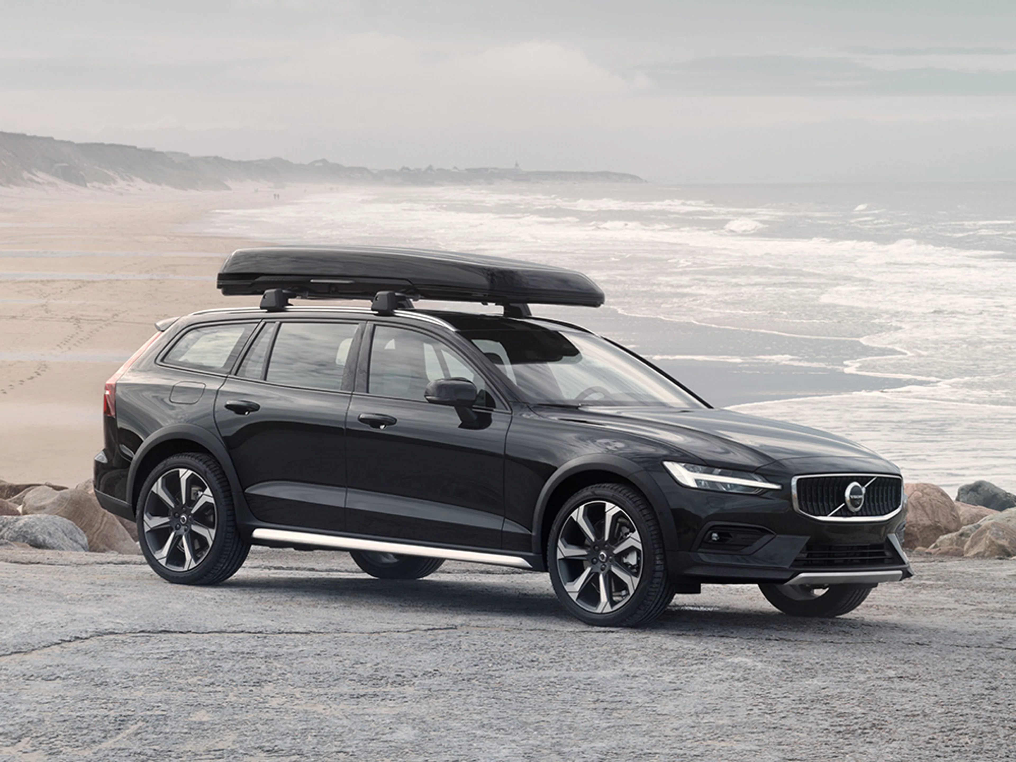 Een zwarte Volvo V60 Cross Country met dakkoffer, geparkeerd aan de kust.