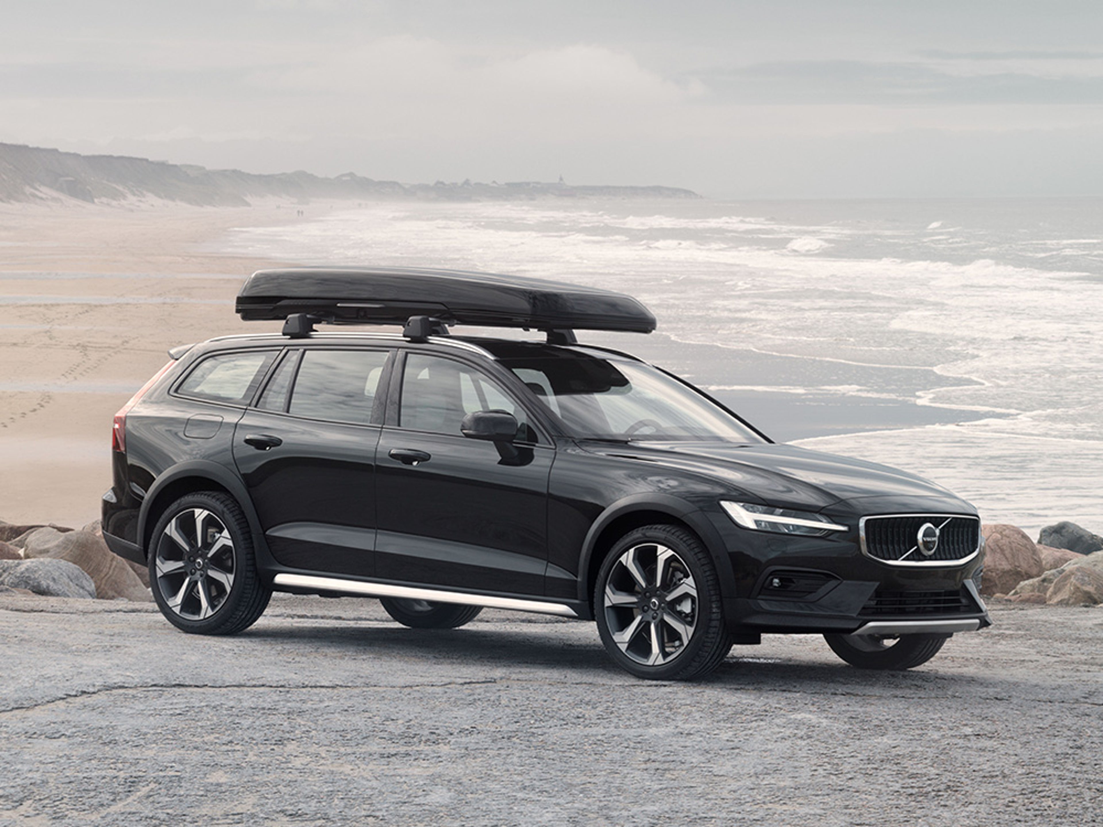 Une Volvo V60 Cross Country noire avec un coffre de toit, garée sur la côte.