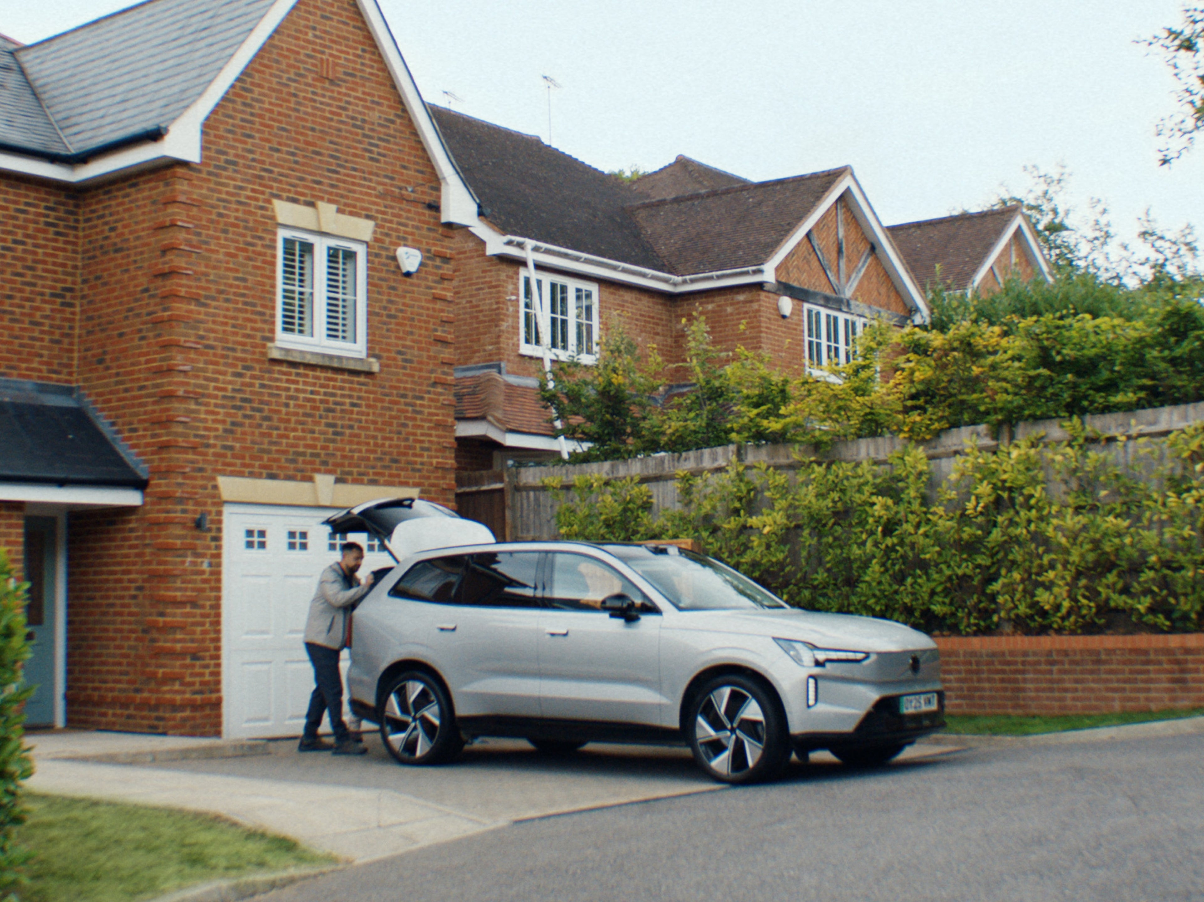 Side view of Volvo EX90 parked on driveway, while someone puts items into the open boot.