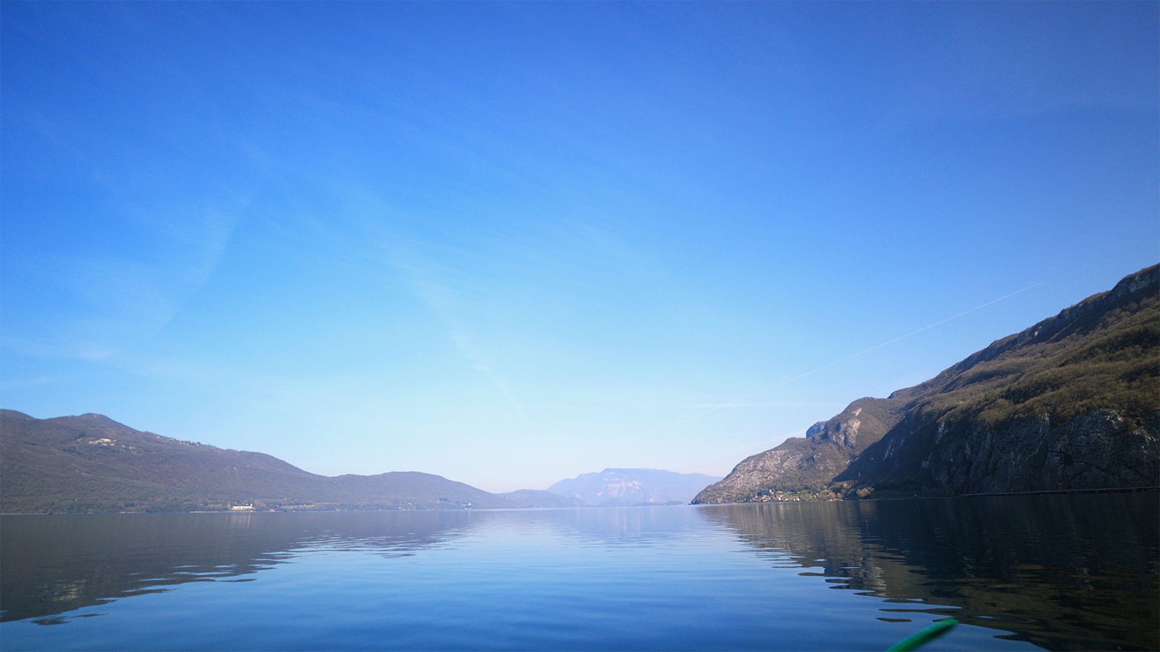Lac calme entouré de montagnes sous ciel bleu, reflet du paysage sur l’eau.