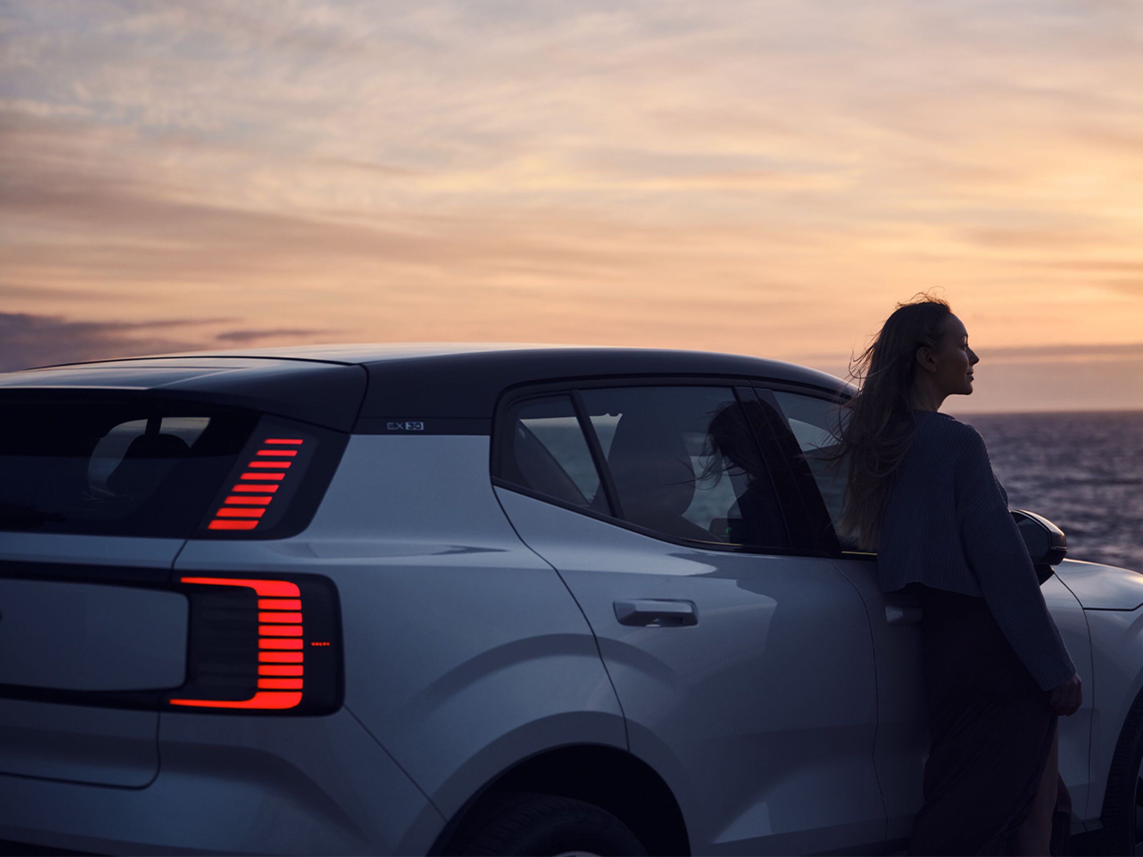 Kvinna vid en Volvo parkerad vid havet i en solnedgång.