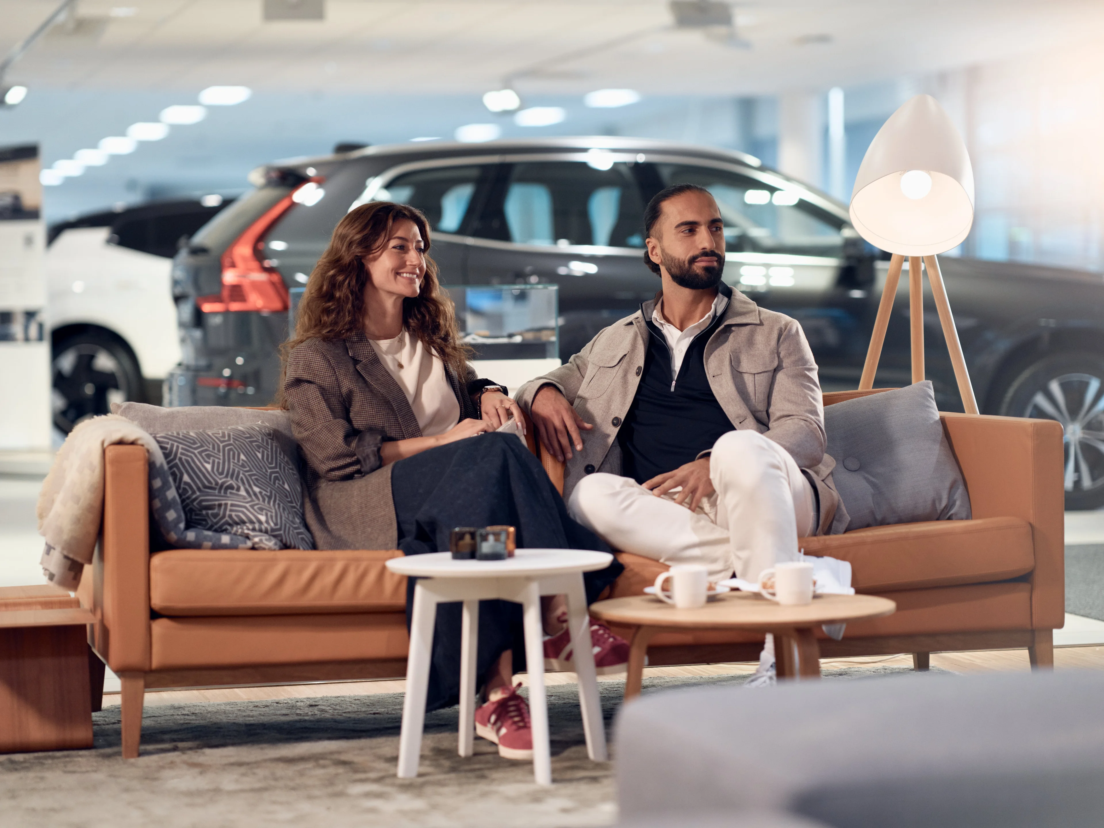 Ein Mann und eine Frau sitzen auf einer braunen Ledercouch in einem Wartebereich. Hinter Ihnen sieht man zwei Volvo Fahrzeuge hinter einer Scheibe stehen.