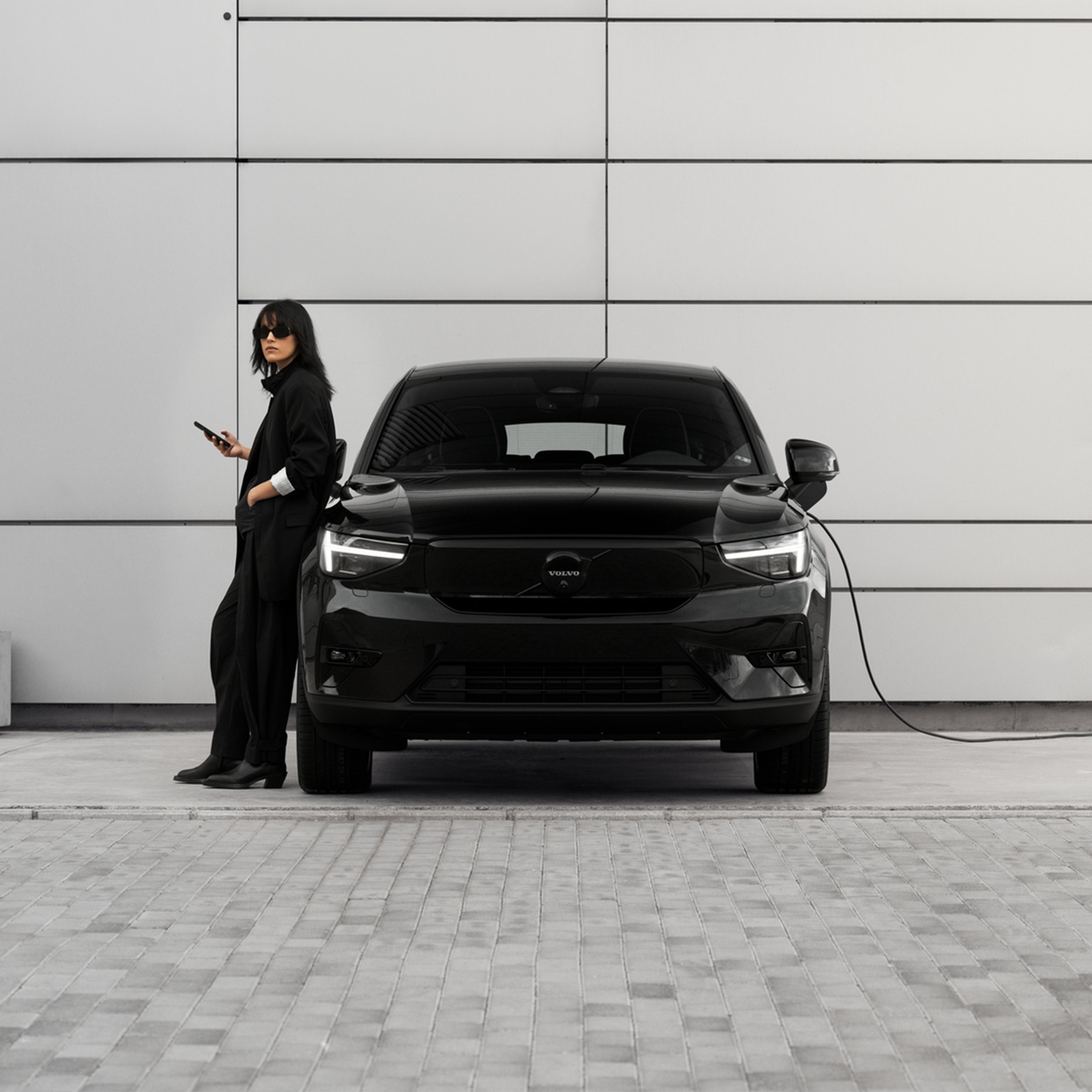 Mustiin pukeutunut nainen nojaa lataukseen kytkettyyn Volvo EC40 täyssähköautoon.