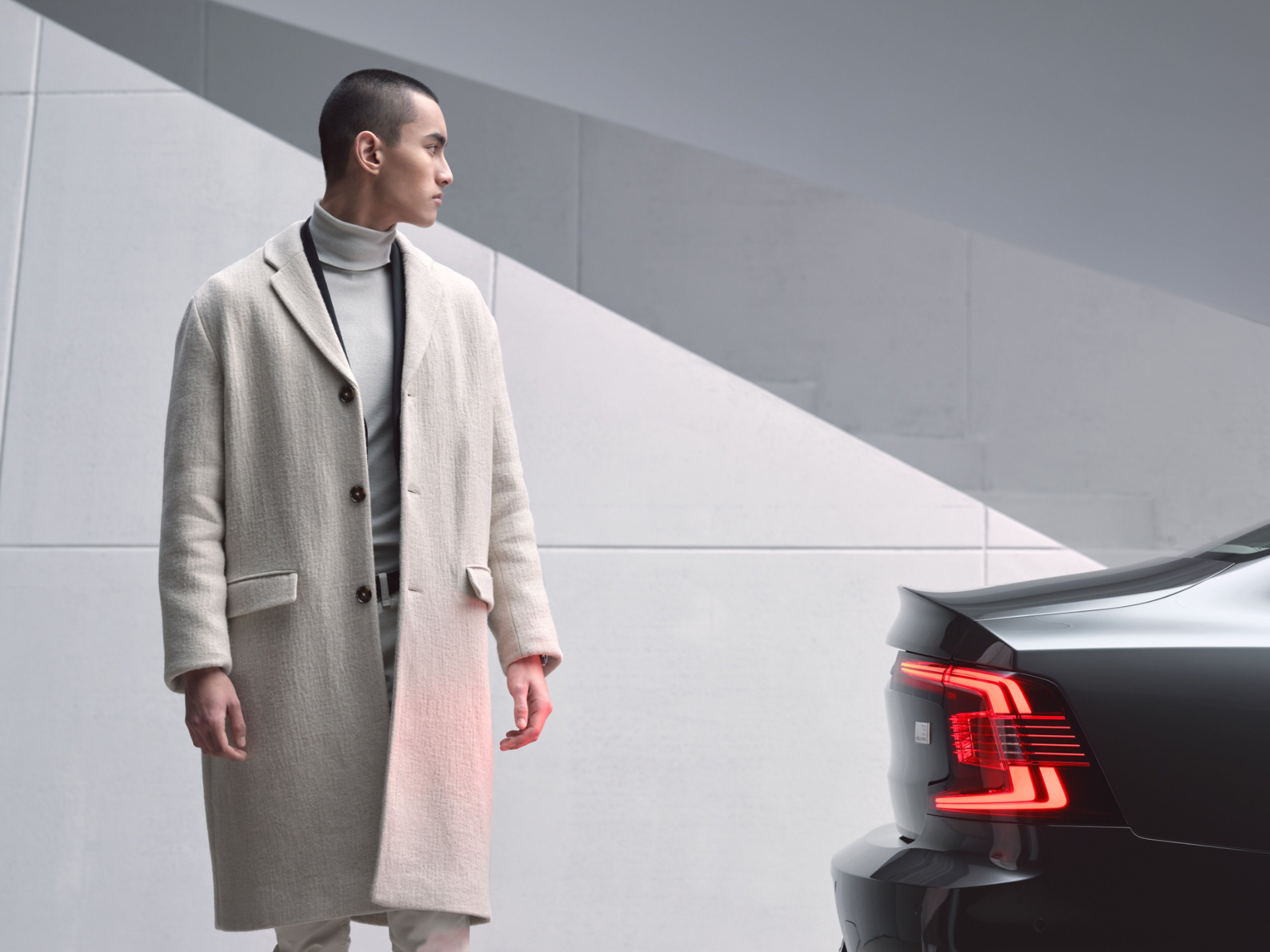 Ein Mann der einen Mantel trägt geht hinter einem Volvo entlang.