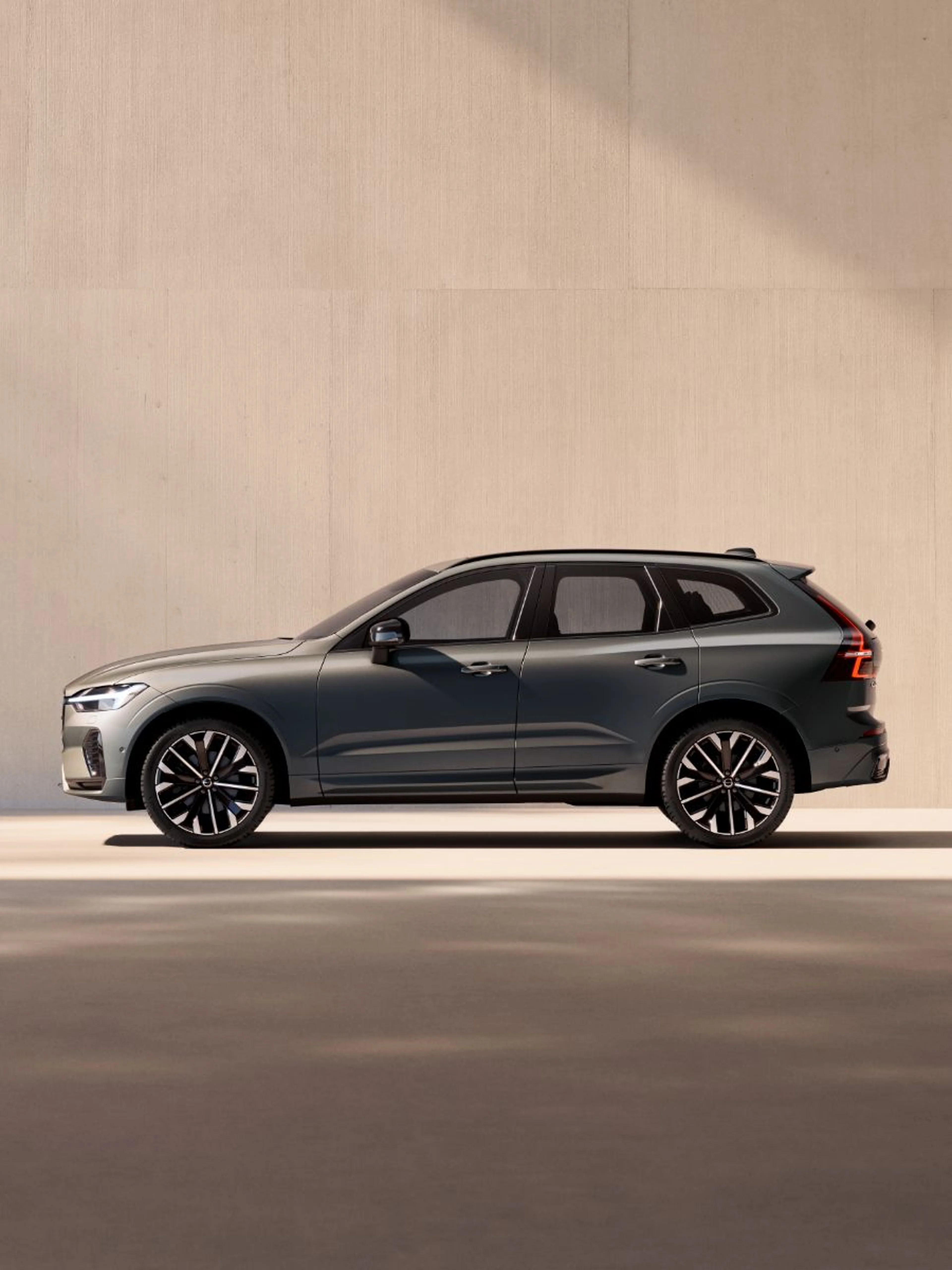 Temno sivi Volvo XC60 prikazan iz bočnega profila, parkiran pred svetlim betonskim zidom v studijskem okolju.