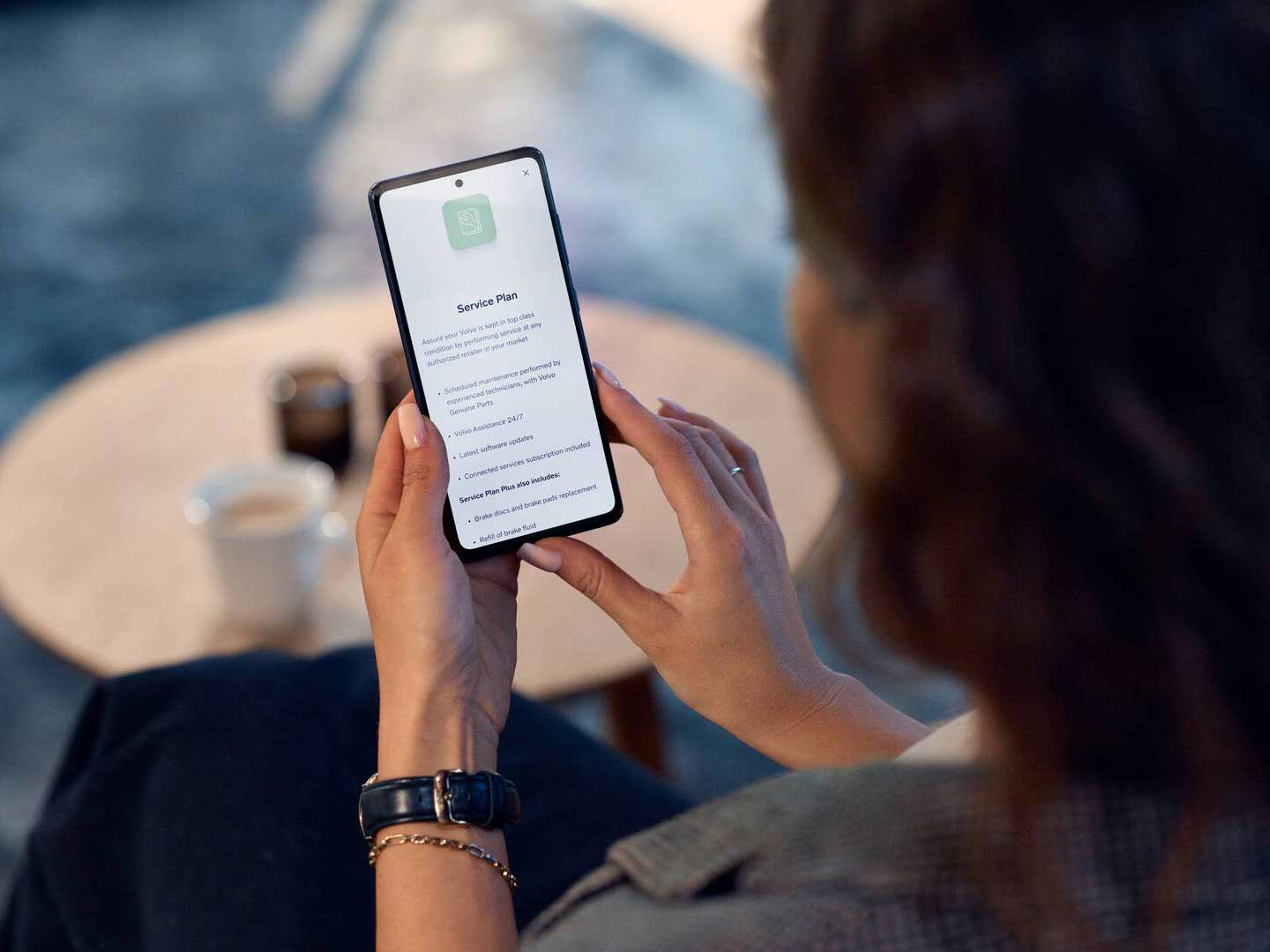 En kvinde ses siddende bagfra med fokus på hendes smartphone, som er logget på Volvo Cars appen og tilgår Service Plan siden.