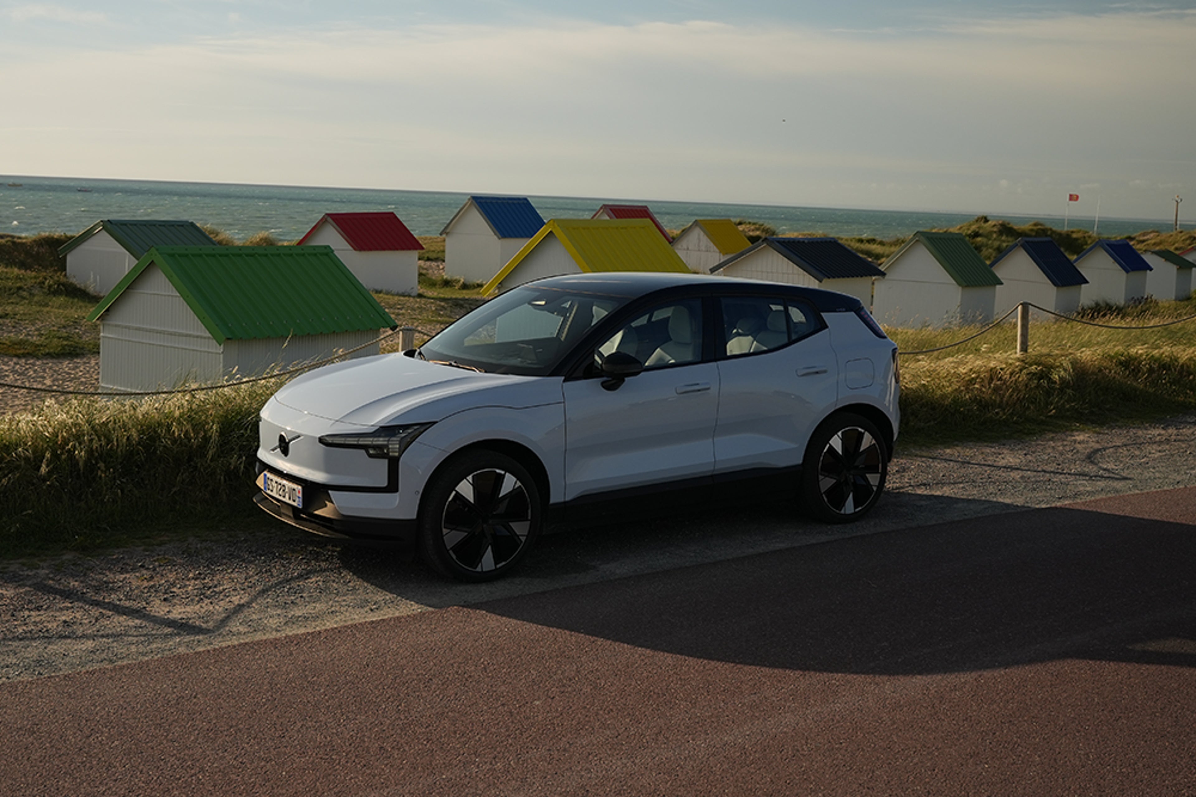 Volvo blanche près de cabanes colorées en bord de mer, ciel bleu et océan en arrière-plan.