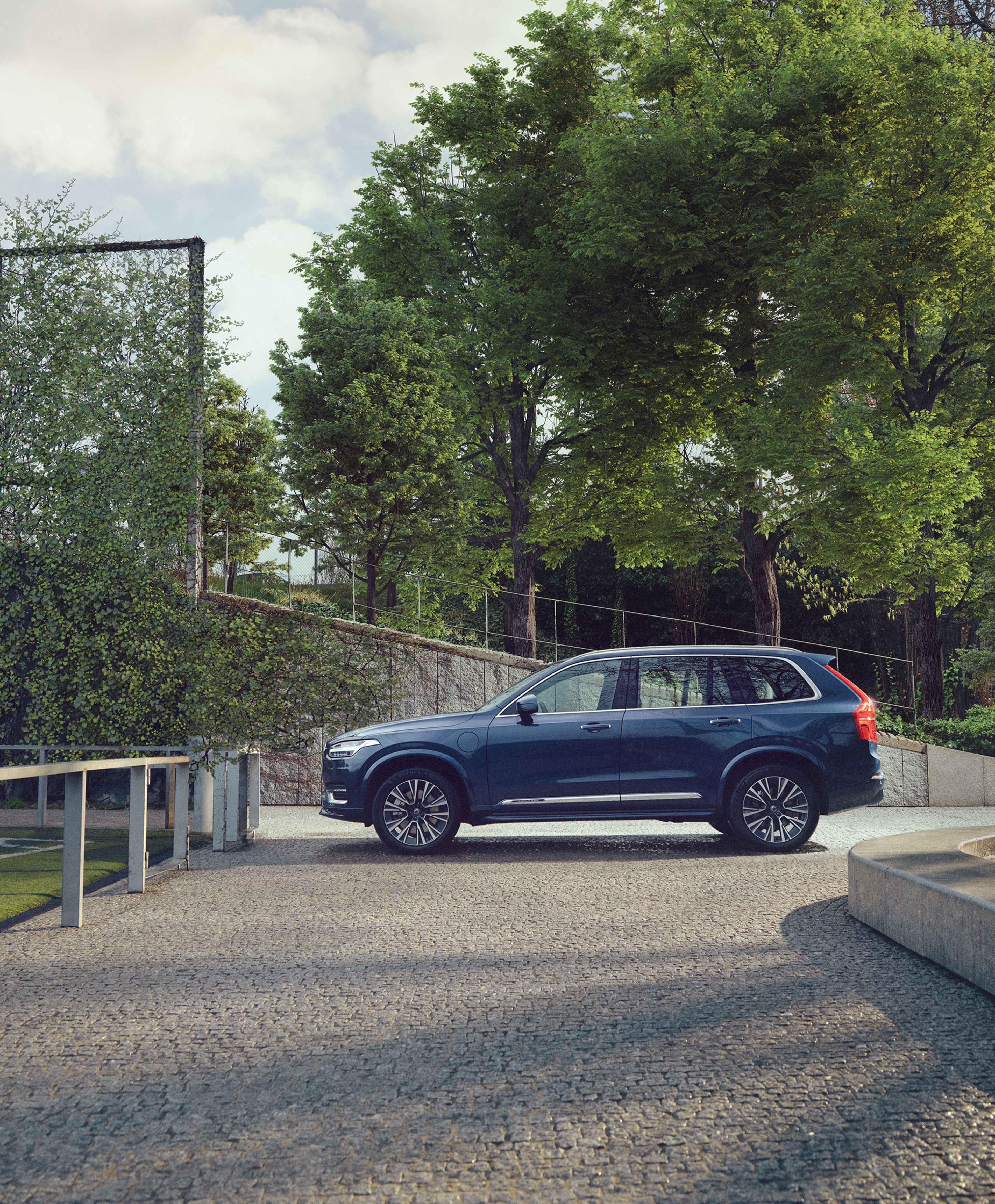 En elegant blå Volvo SUV holder parkeret på en brostensbelagt indkørsel omgivet af frodige grønne træer. Den rolige atmosfære udstråler en følelse af ro og luksus.