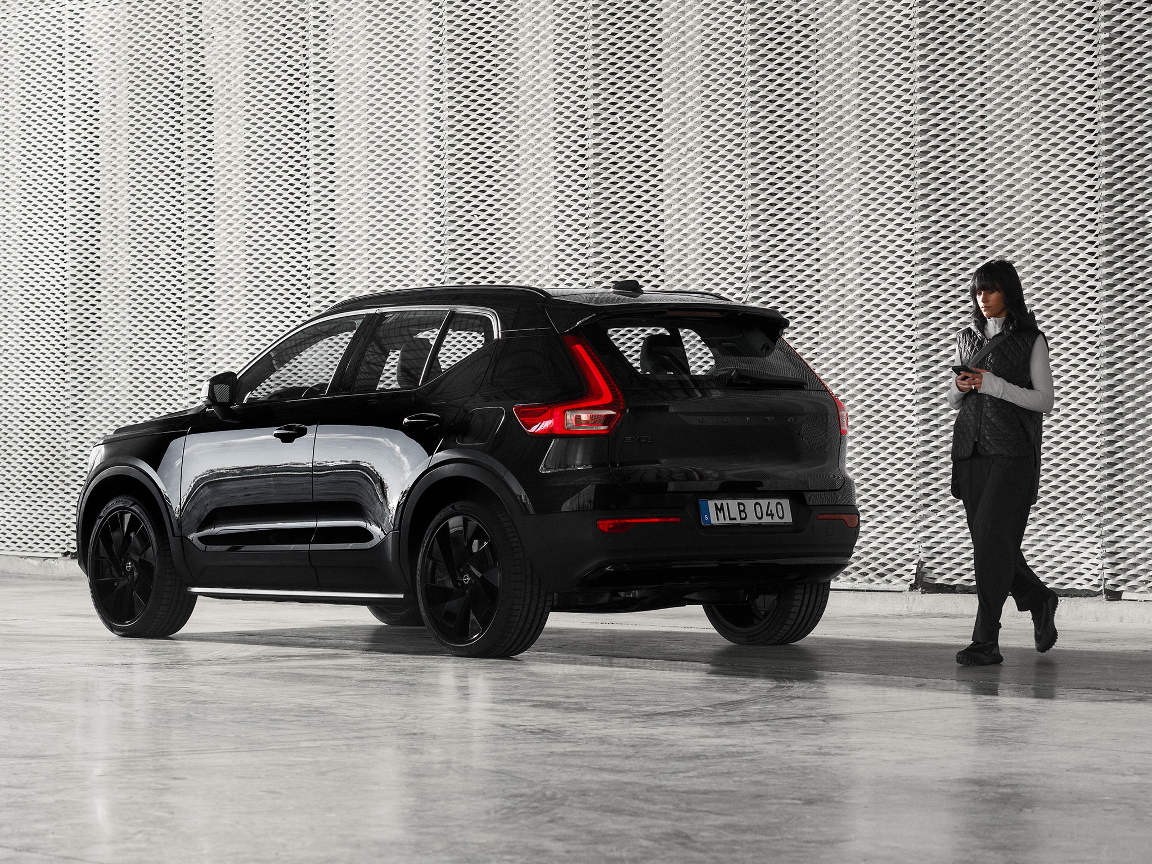 Mustiin vaatteisiin pukeutunut nainen kävelee puhelin kädessään EX40 Black Edition täyssähköauton takaa.
