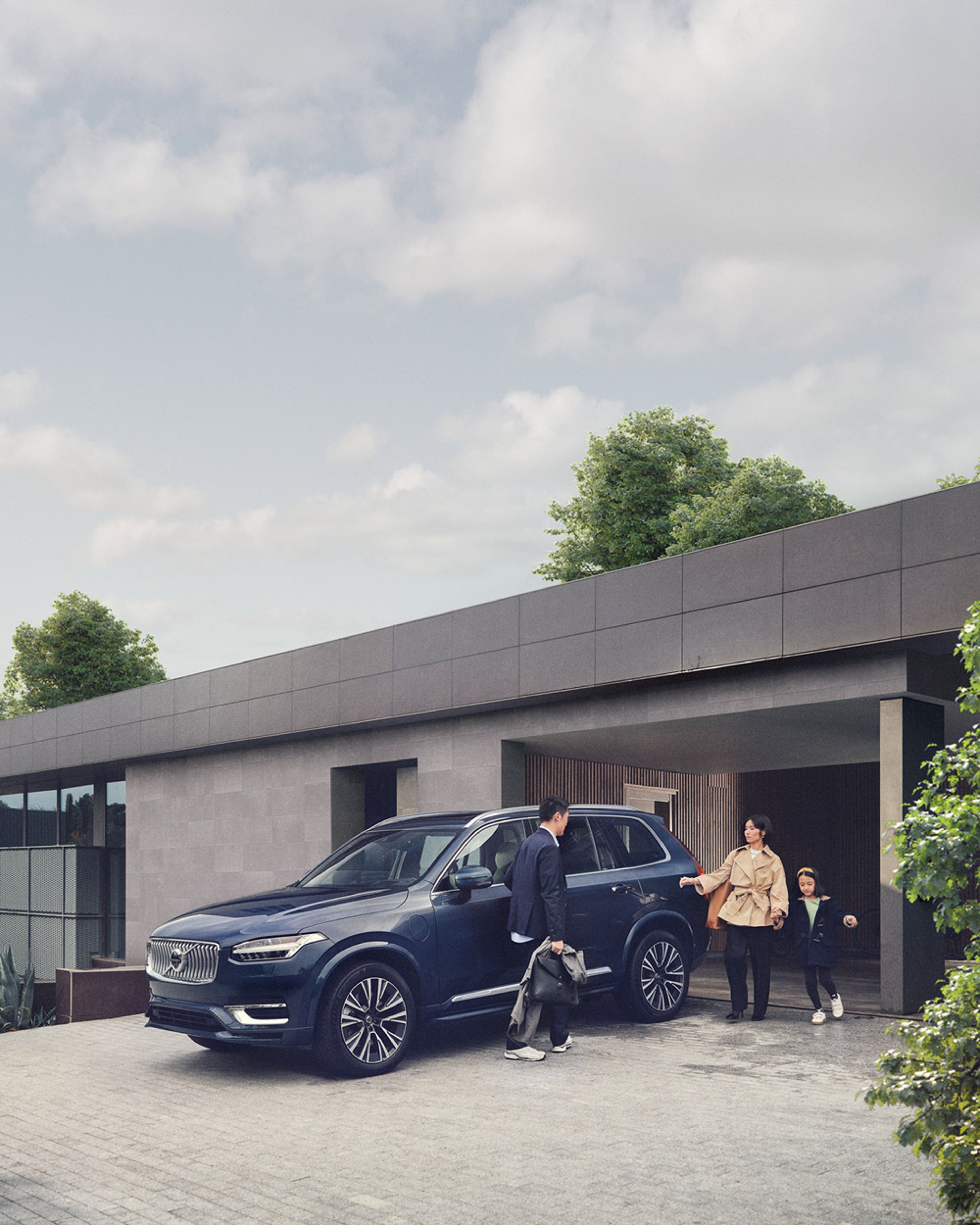 Une famille descend d'un SUV Volvo bleu foncé devant une maison moderne à toit plat. Le père sort de la voiture, tandis que la mère et la fille sont déjà sorties et se dirigent vers l'entrée de la maison.