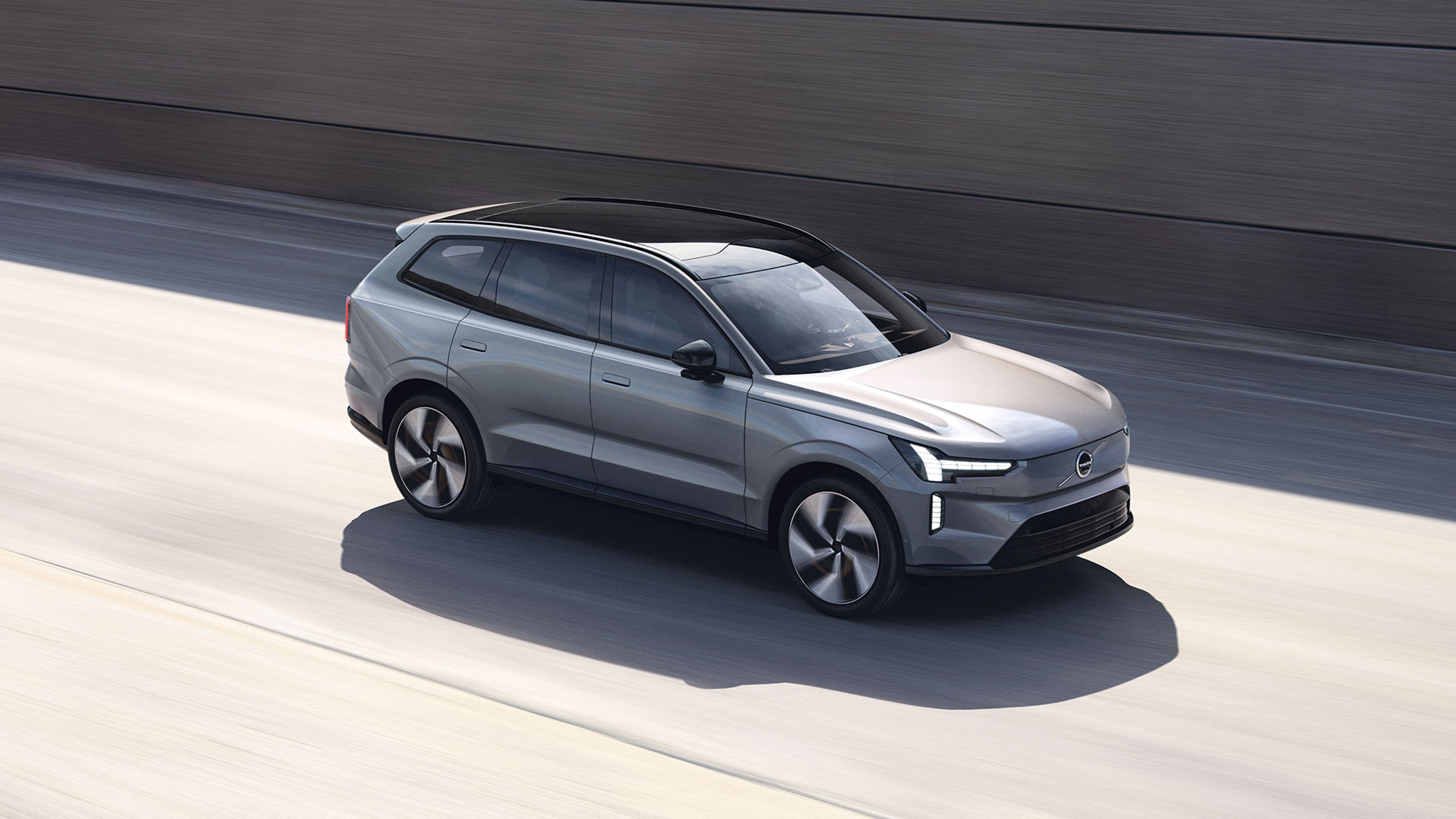 Volvo EX90 gris roulant sur une route ensoleillée avec un mur en arrière-plan